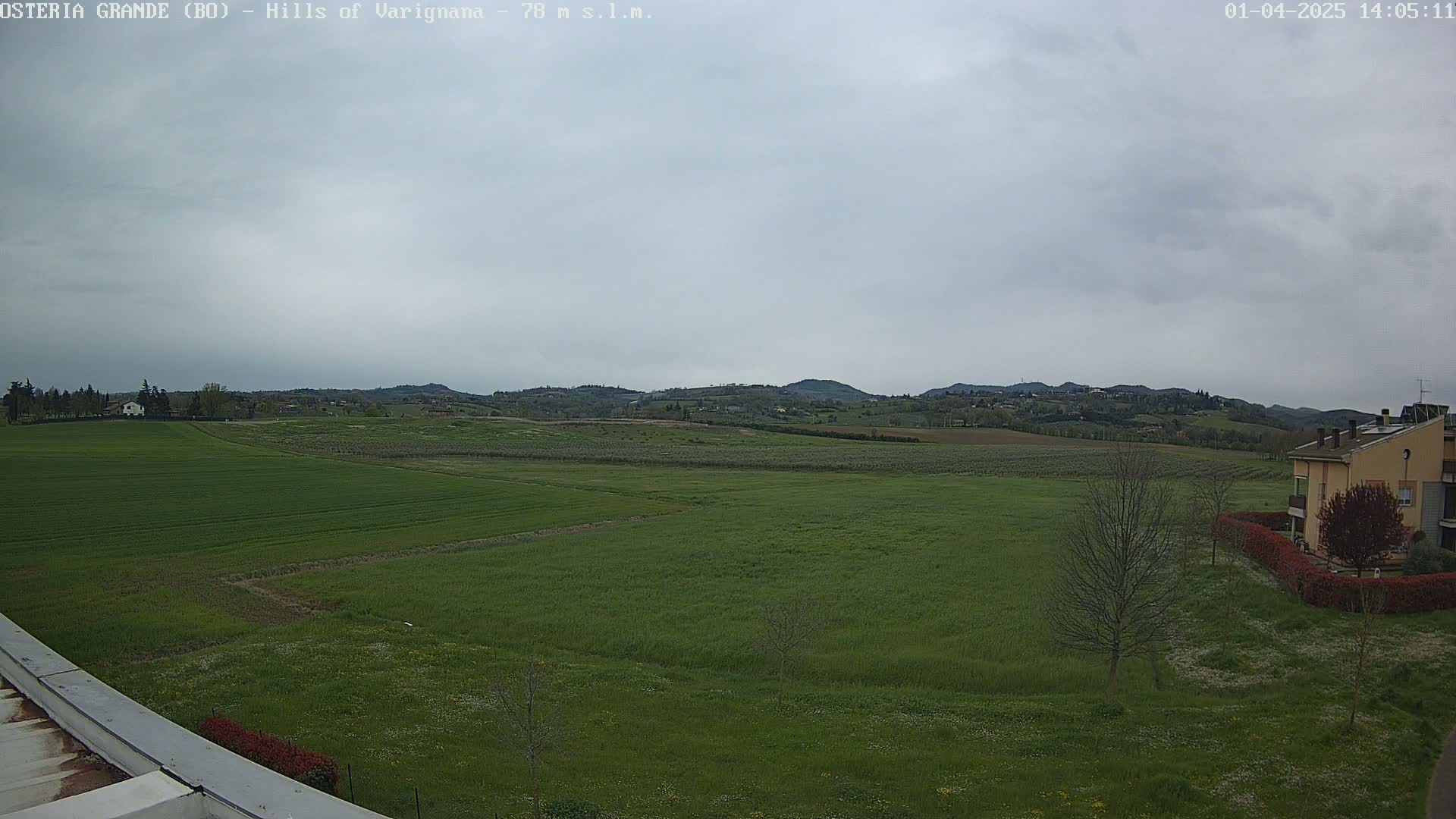 A wide, verdant field stretches towards a line of rolling hills under a mostly cloudy sky.