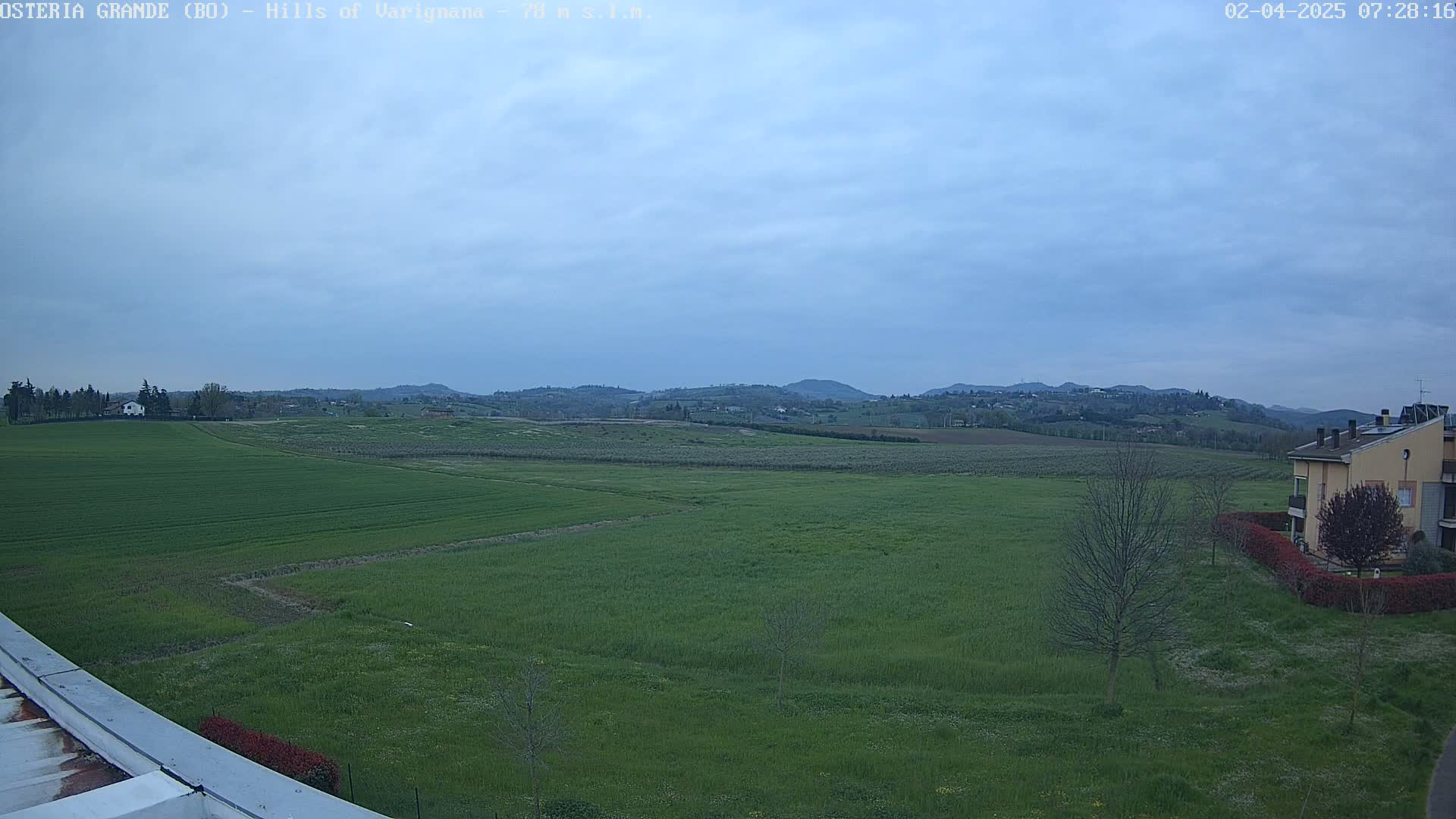 A sprawling green field stretches out to rolling hills under a mostly cloudy, light-blue sky.