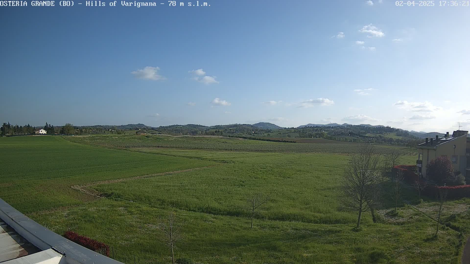 A mostly sunny day reveals expansive green fields and rolling hills in the distance, with a few buildings visible near the bottom right.