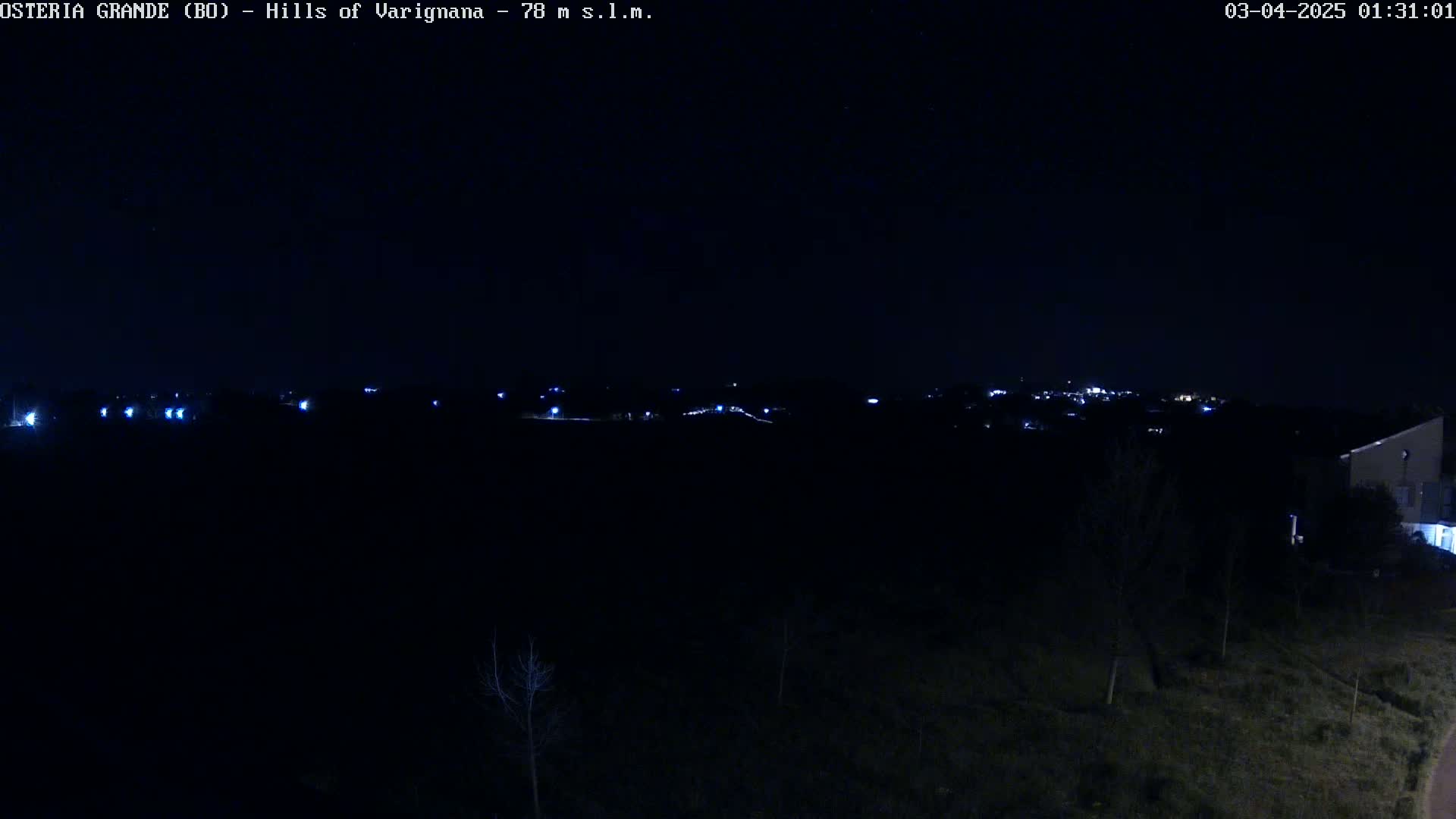 A nighttime, long shot of a dark landscape with a few faint lights in the distance and a building partially visible in the bottom right corner.