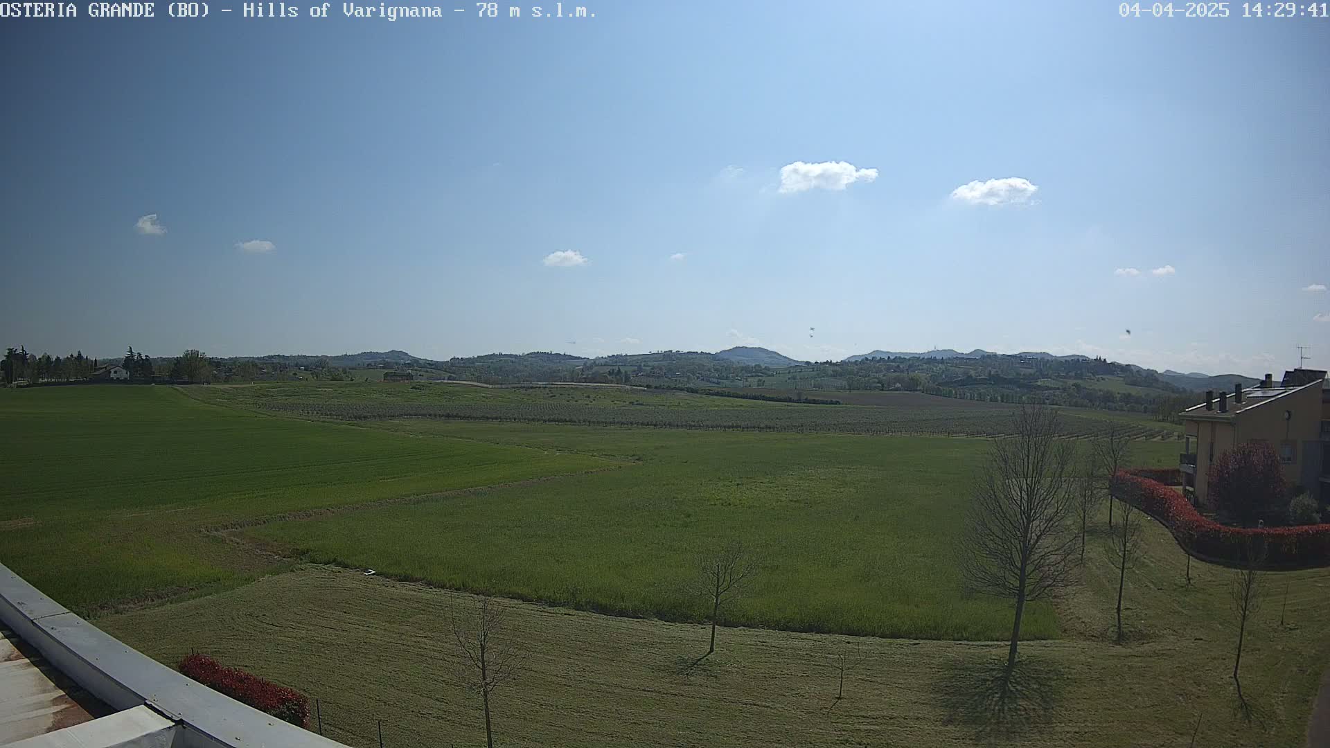 A sunny day reveals a sprawling landscape of green fields, dotted with a few trees and a building in the distance, all set against a backdrop of rolling hills under a mostly clear sky.