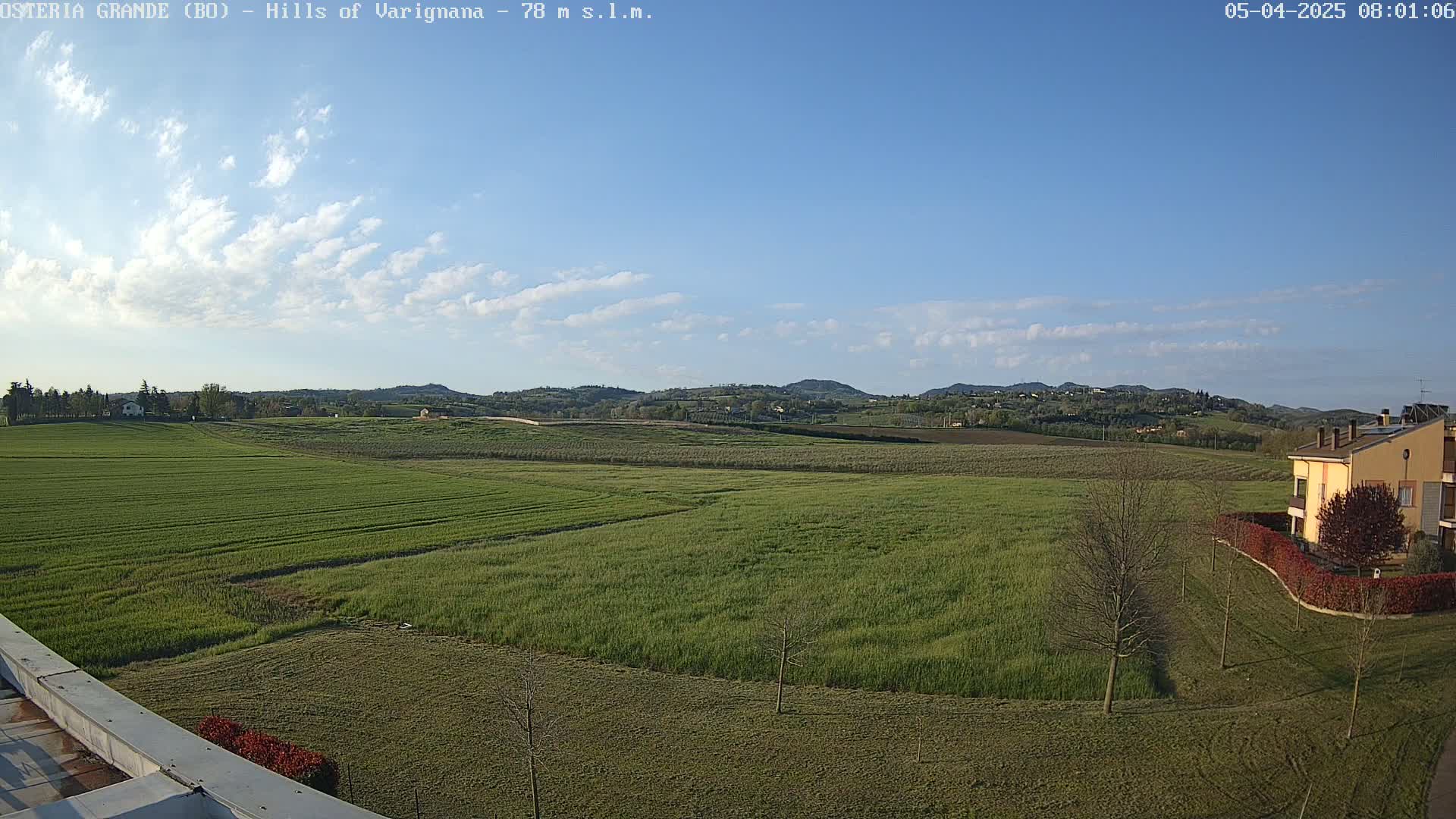 A sprawling green field stretches out beneath a mostly clear blue sky dotted with a few clouds, with distant hills and a few buildings visible in the background.