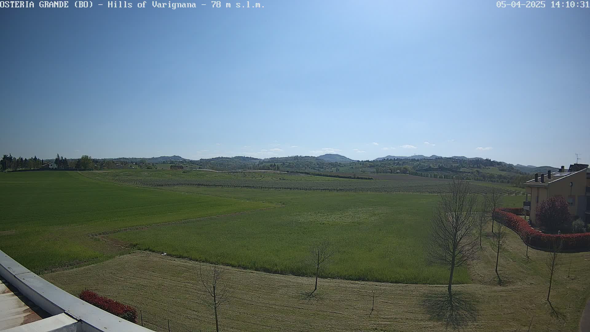 A sunny day reveals a sprawling green landscape of fields and orchards, rolling hills in the distance, and a building with a hedge to its right, near the lower right corner.