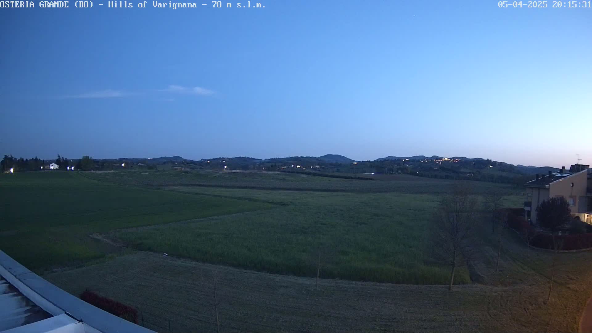A twilight view shows rolling green hills and fields under a clear, dusky blue sky, with a few buildings visible in the foreground and distance.