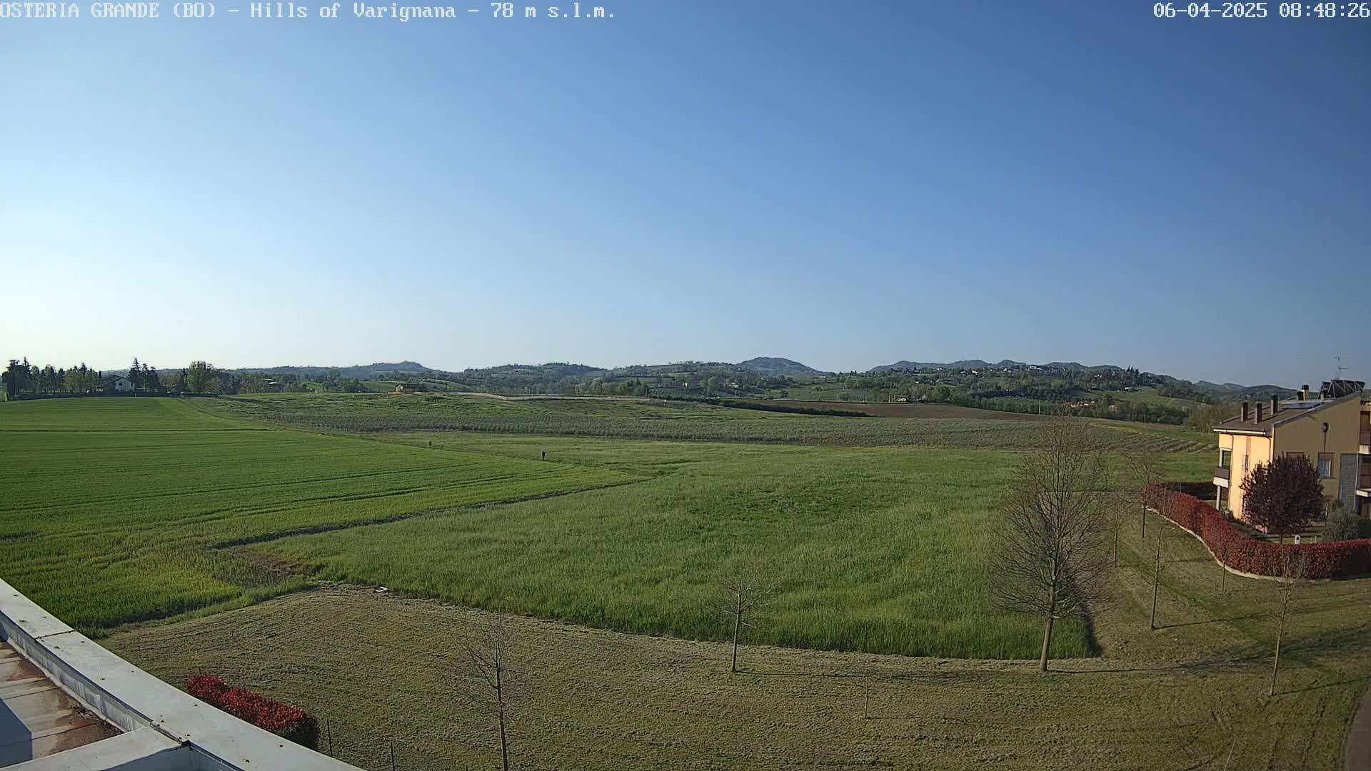 A wide, expansive view of green fields under a clear blue sky shows a few buildings in the distance and a few trees nearby.