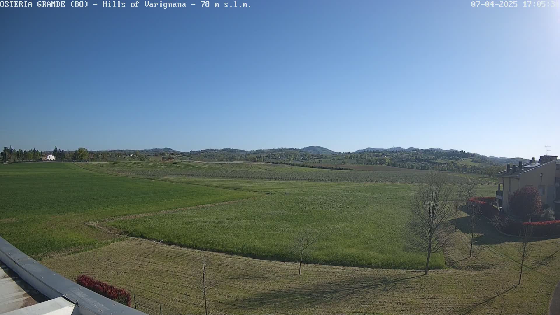 A wide, sunny outdoor shot shows rolling green hills and fields, with a few buildings visible in the foreground and distant background under a clear blue sky.