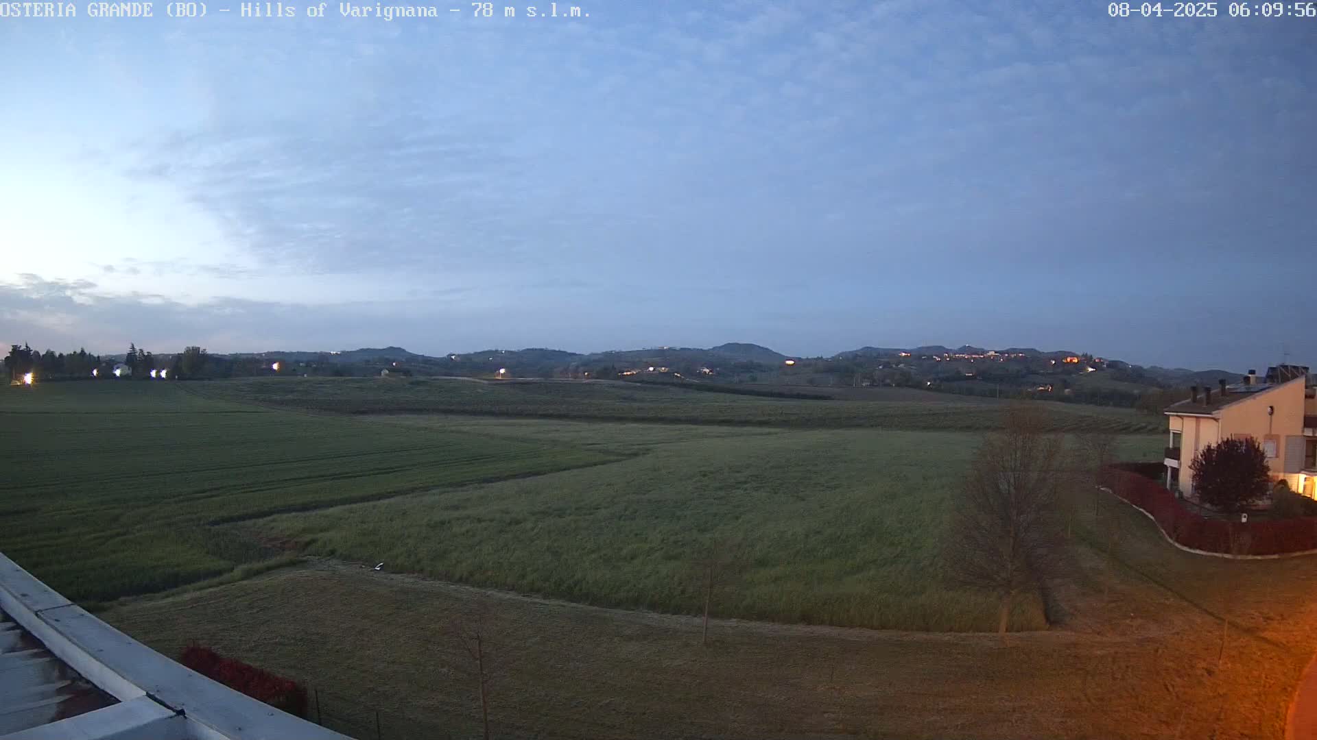 A wide, flat field of green grass and farmland stretches out to distant, dimly lit hills under a twilight sky.