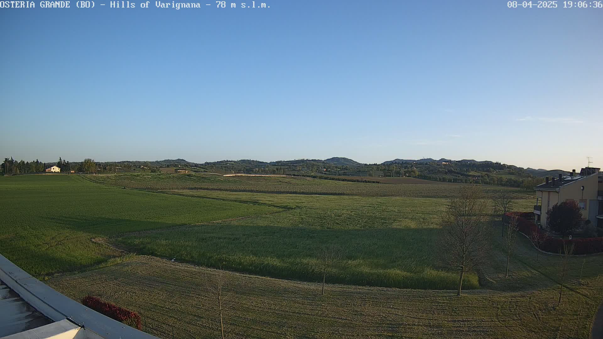 Under a clear blue sky, a vast expanse of green fields stretches out to rolling hills in the distance, with a few buildings visible near the bottom right.