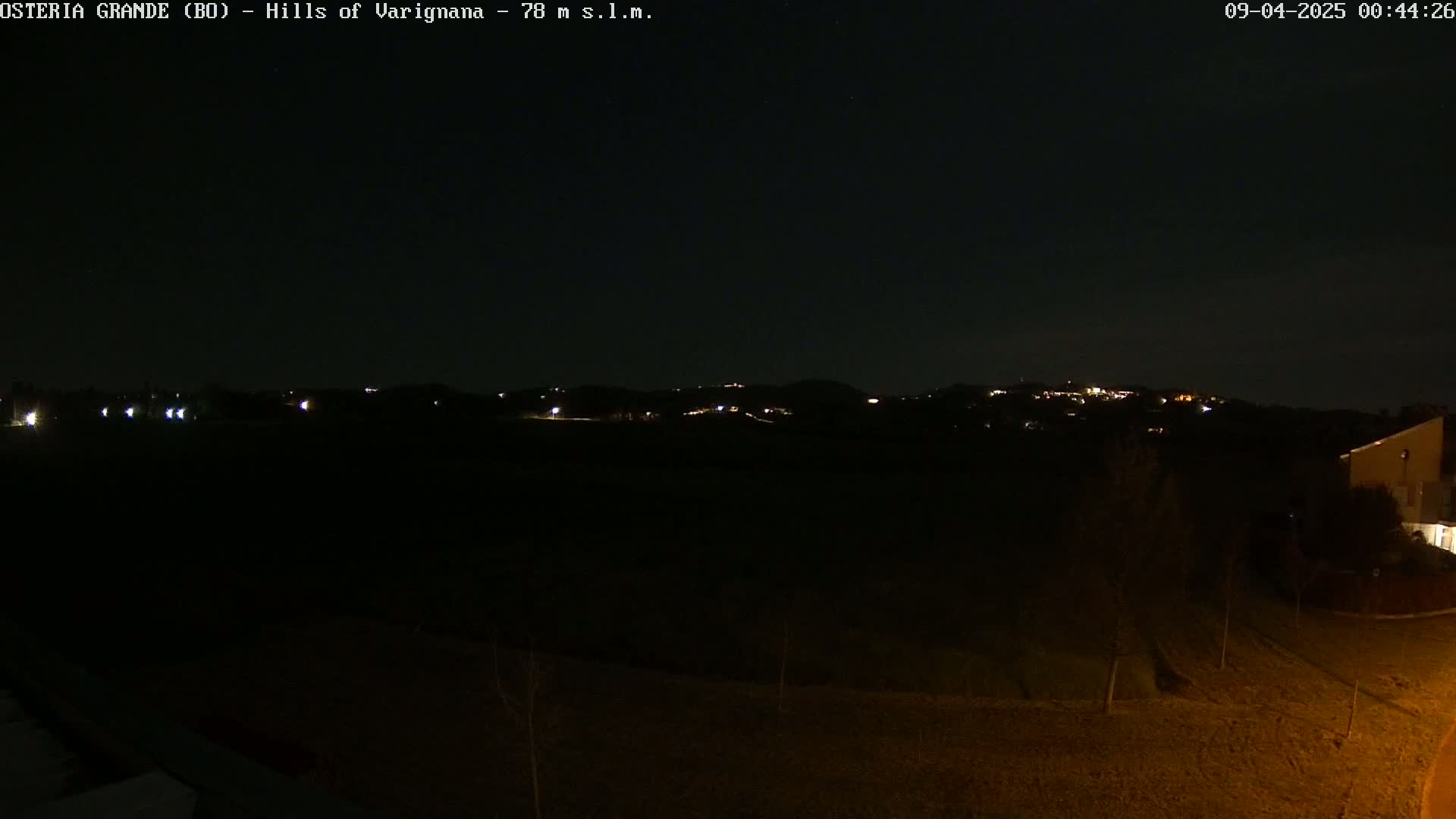 A nighttime, low-light view of dimly lit hills in the distance, with a dark foreground containing sparsely vegetated land and a portion of a building.