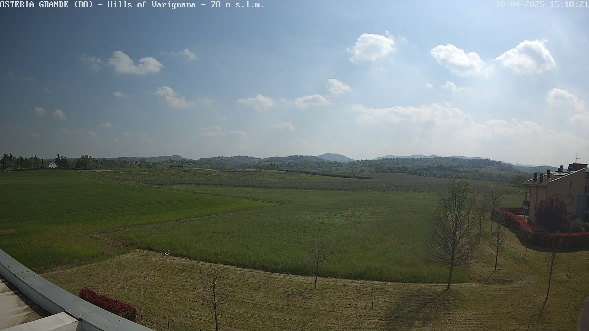 A partly cloudy sky overlooks a vast expanse of green fields, punctuated by a few buildings and trees in the foreground and rolling hills in the distance.