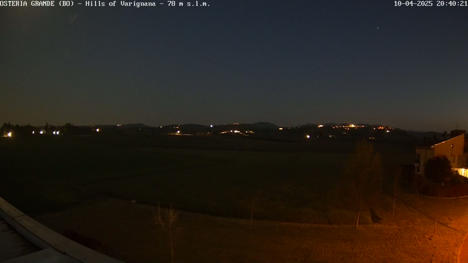 A nighttime view of dimly lit hills in the distance, a flat field in the foreground, and a few buildings to the right, under a dark, clear sky.