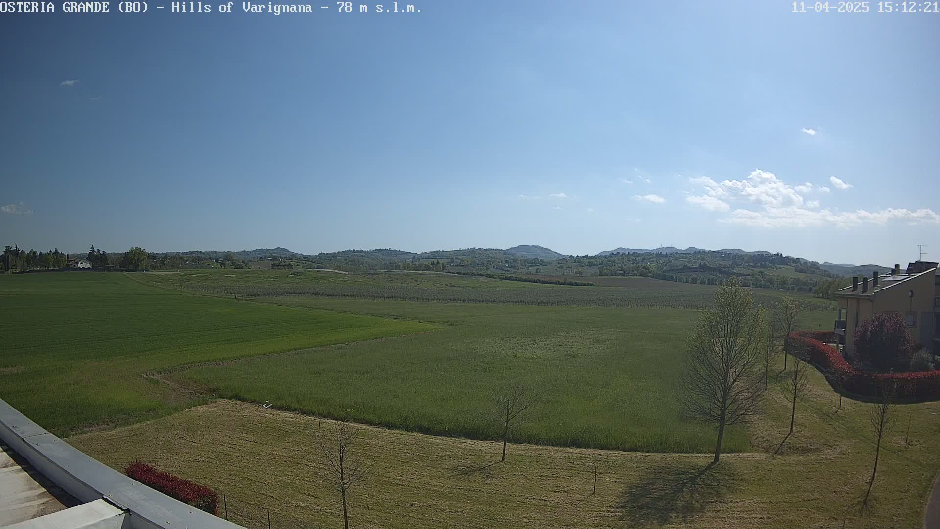A mostly sunny day reveals a sprawling landscape of green fields and distant rolling hills, with a few trees and a building visible in the foreground.