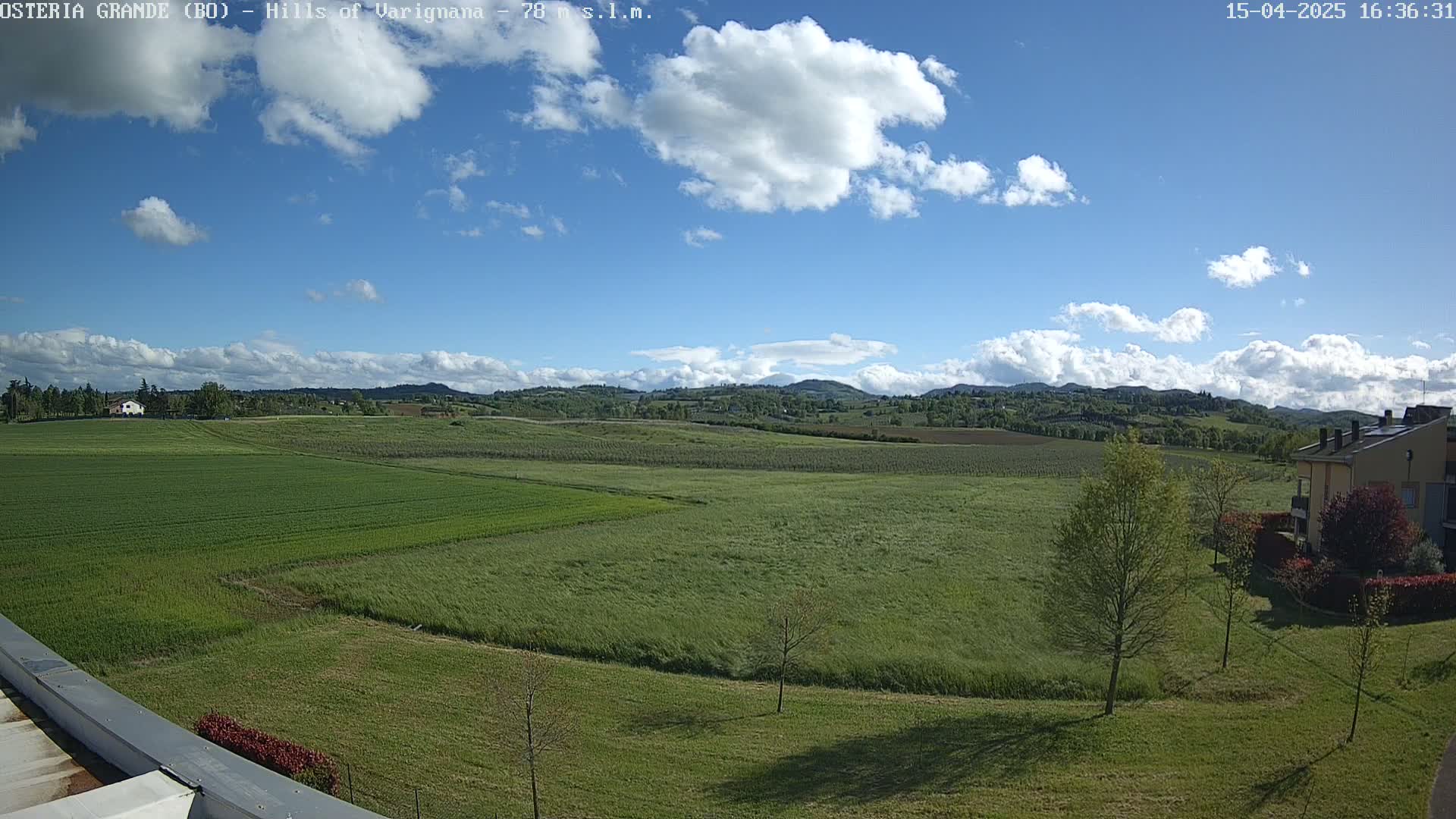 A partly cloudy sky overlooks expansive green fields and low hills, with a few buildings visible in the lower right corner.