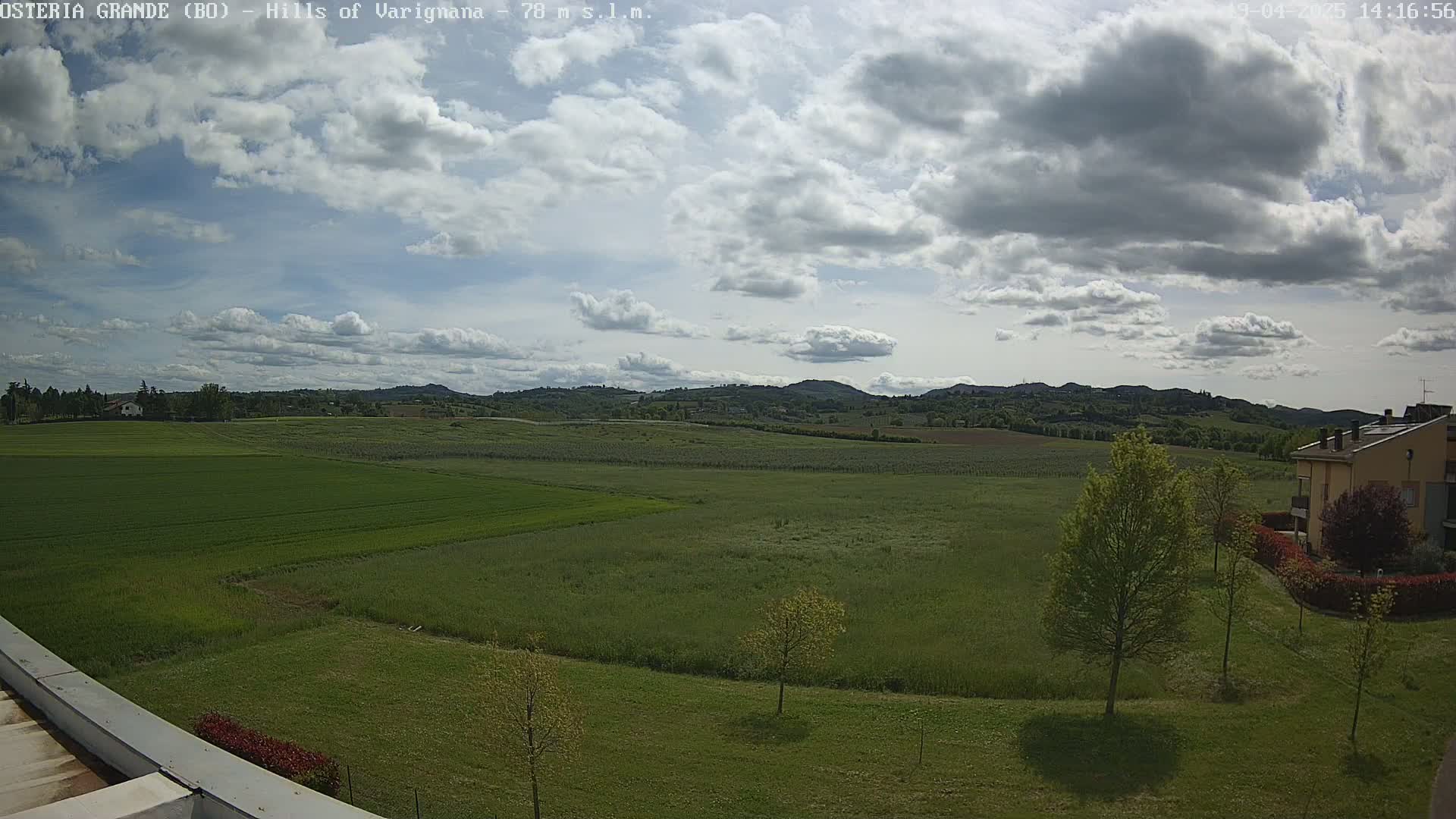 A partly cloudy day reveals a sprawling green landscape of fields and hills, with a few buildings visible in the bottom right.