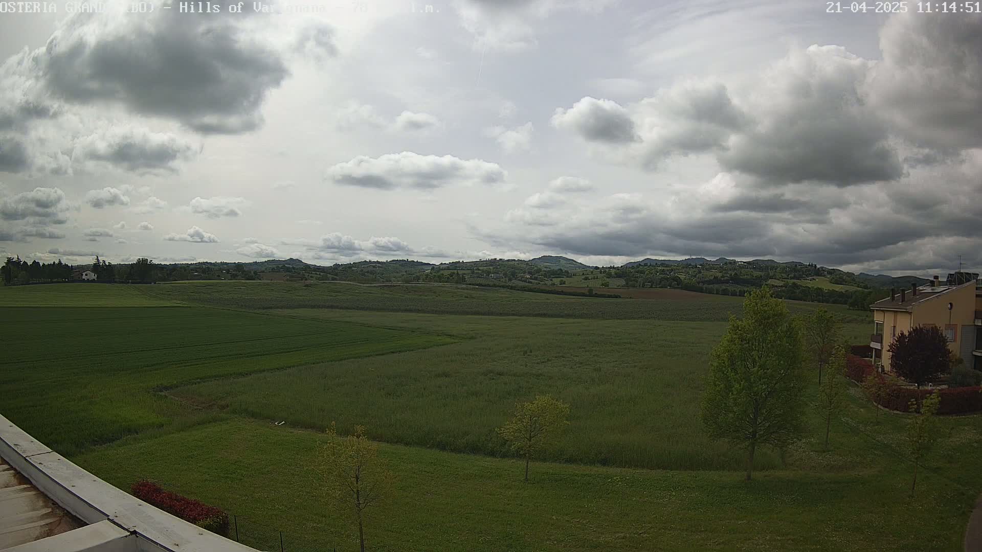 A partly cloudy sky overlooks a vast expanse of green fields and distant hills, with a few buildings visible near the bottom right.
