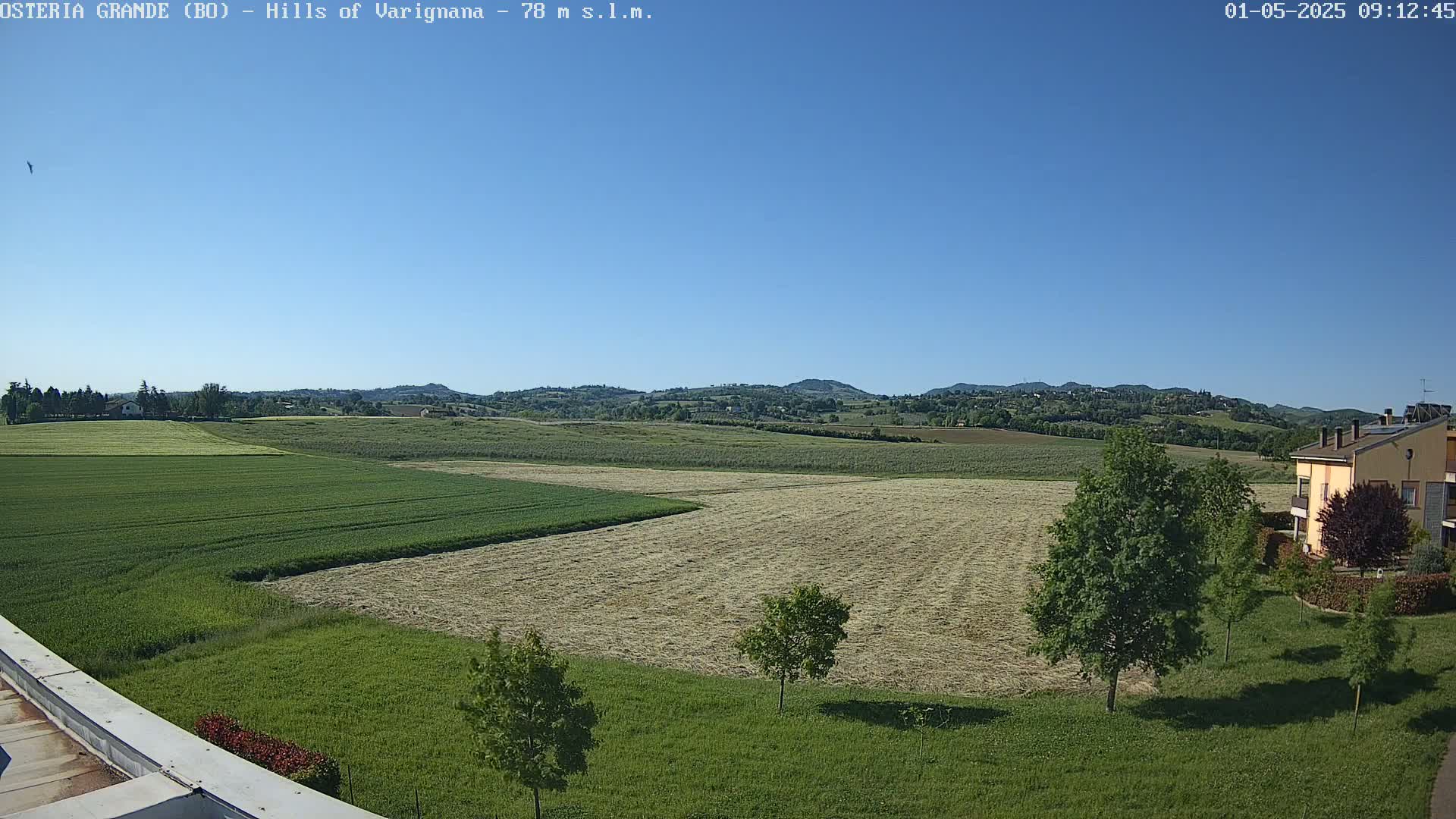 Under a clear blue sky, a vast expanse of green and tan fields stretches towards rolling hills in the distance, with a few buildings and trees visible near the bottom right.