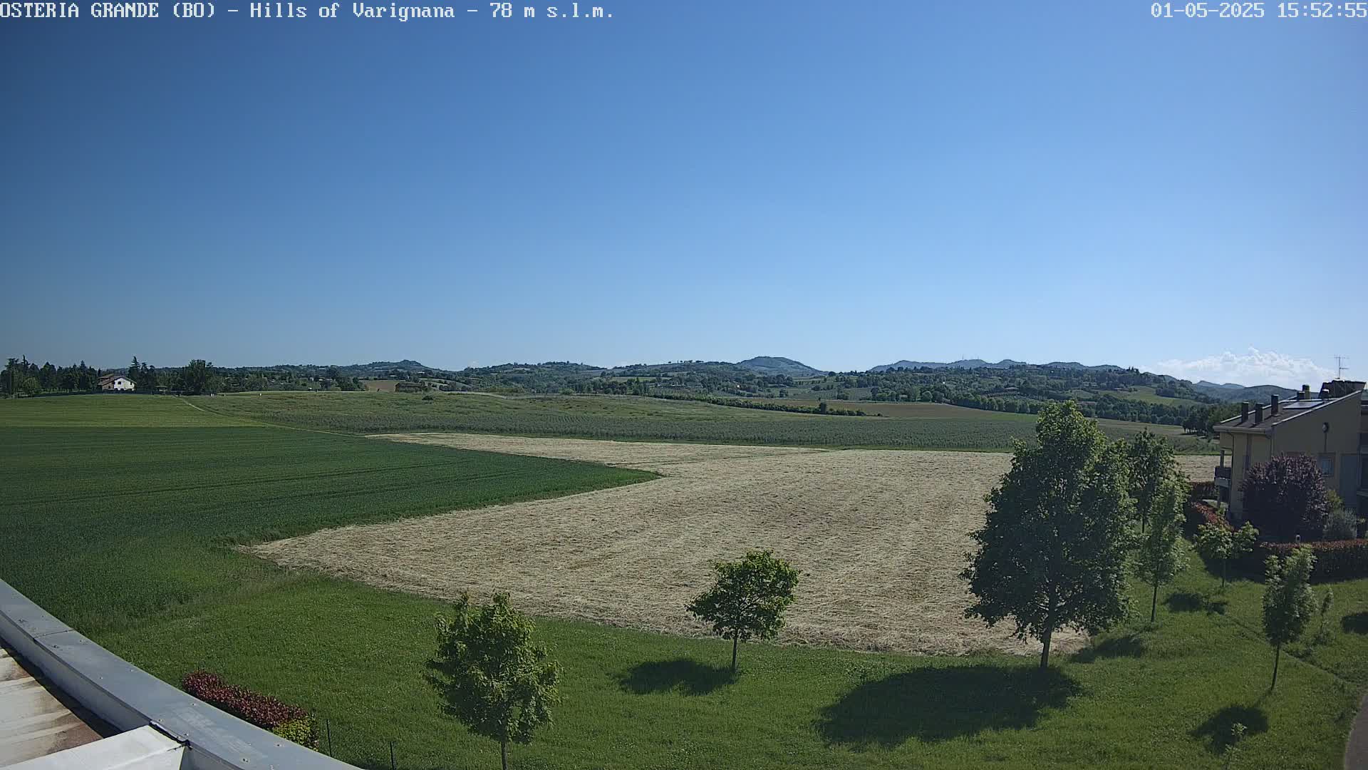 A sunny day reveals rolling green hills dotted with trees and fields of hay, a few buildings visible in the distance.