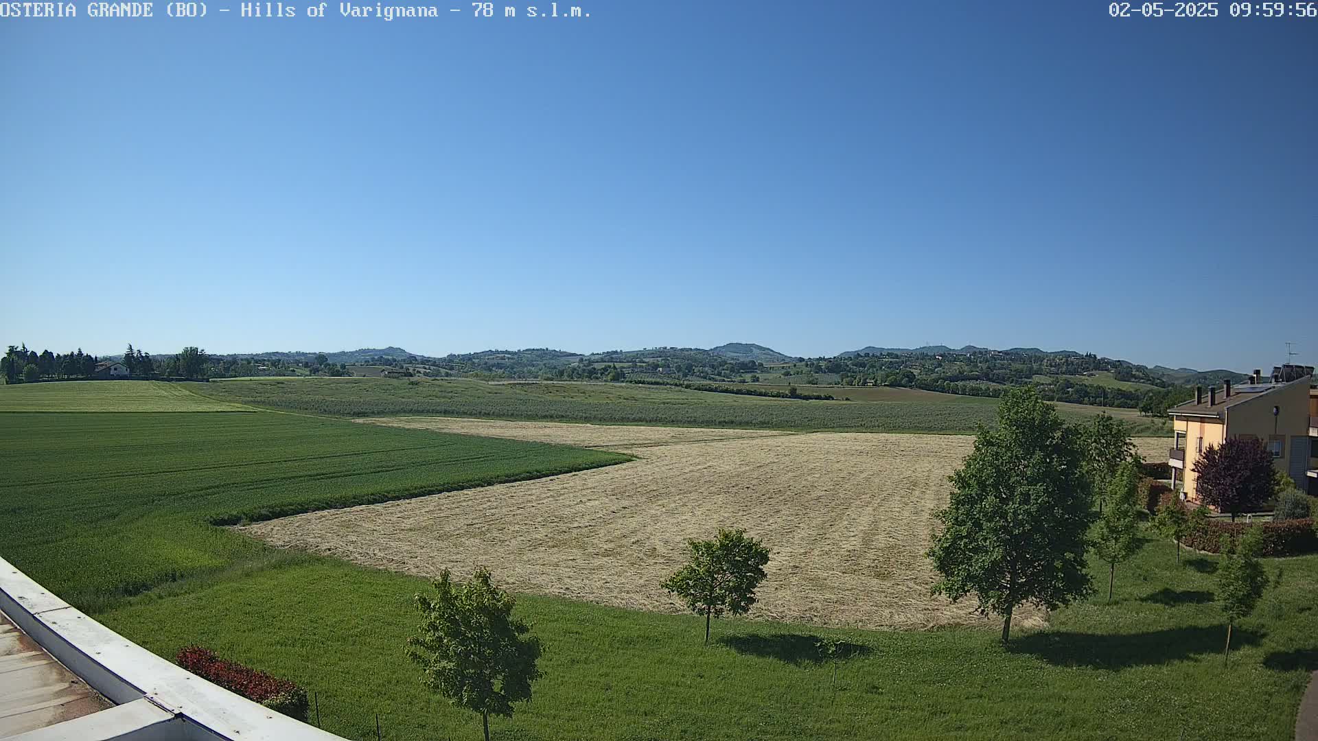 Under a clear blue sky, a view of rolling hills reveals a patchwork of harvested and growing fields, with a few buildings visible in the distance and trees in the foreground.