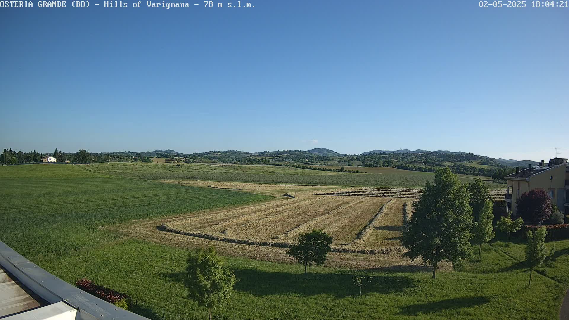 Under a clear blue sky, a vast expanse of farmland displays rows of freshly harvested hay, interspersed with green fields and a few buildings in the distance.