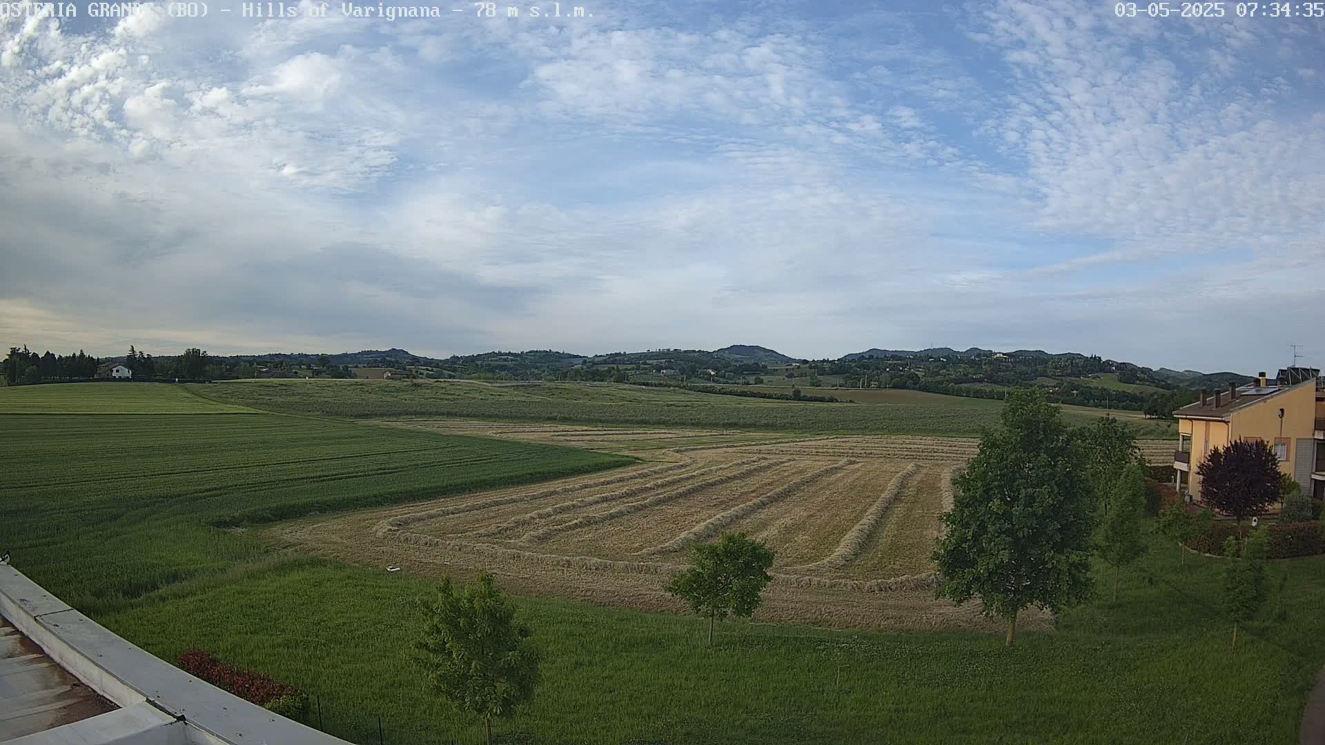 A partly cloudy sky overlooks a landscape of rolling green hills, harvested fields with hay swaths, and a few buildings.