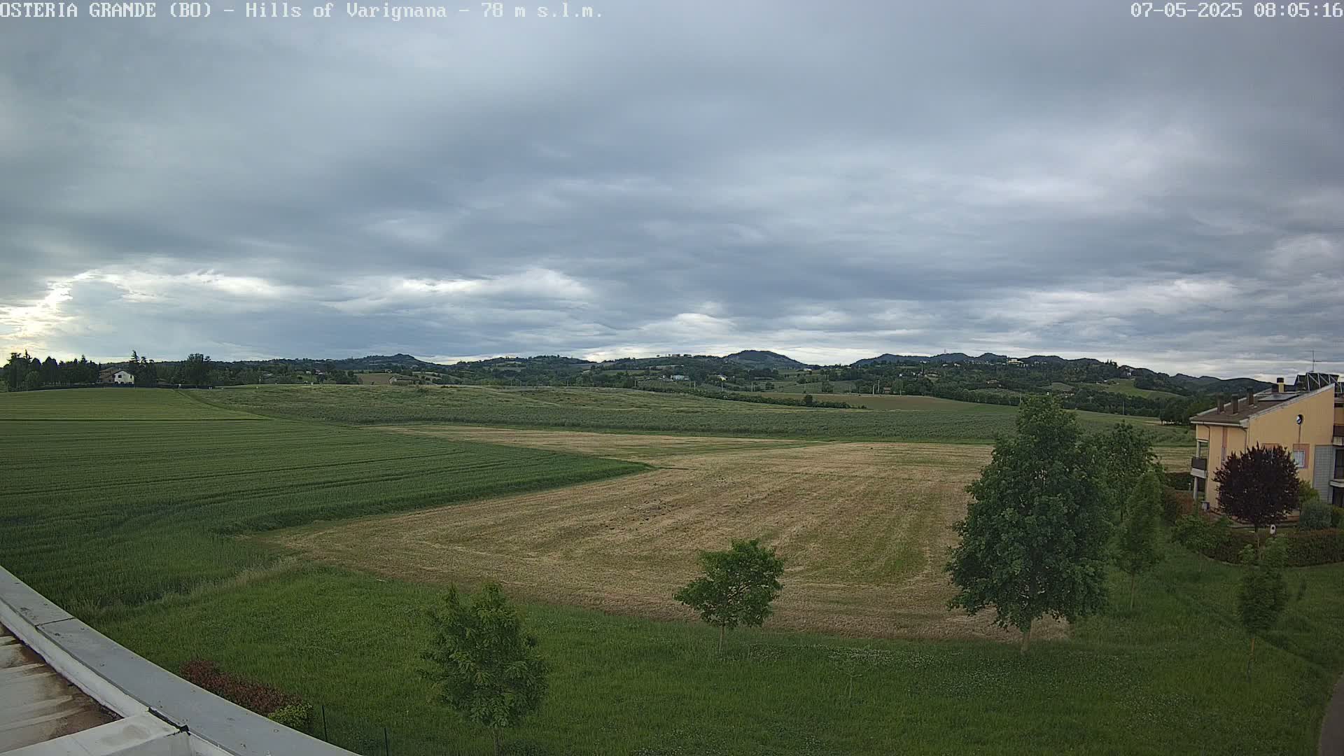 Under a cloudy sky, a view of rolling hills dotted with farm fields, a few houses, and some trees is seen from a slightly elevated vantage point.