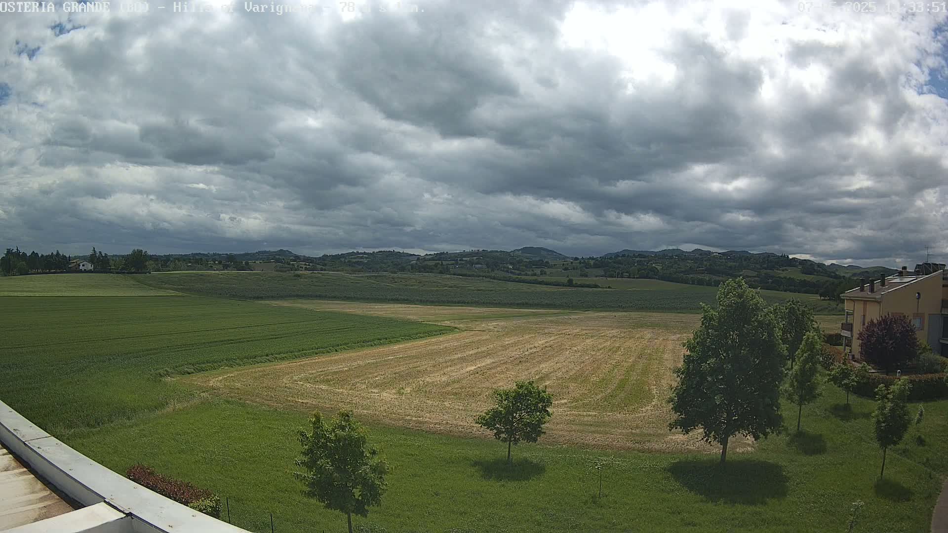 Under a cloudy sky, a wide expanse of farmland with fields of varying shades of green and tan stretches out to rolling hills in the distance.