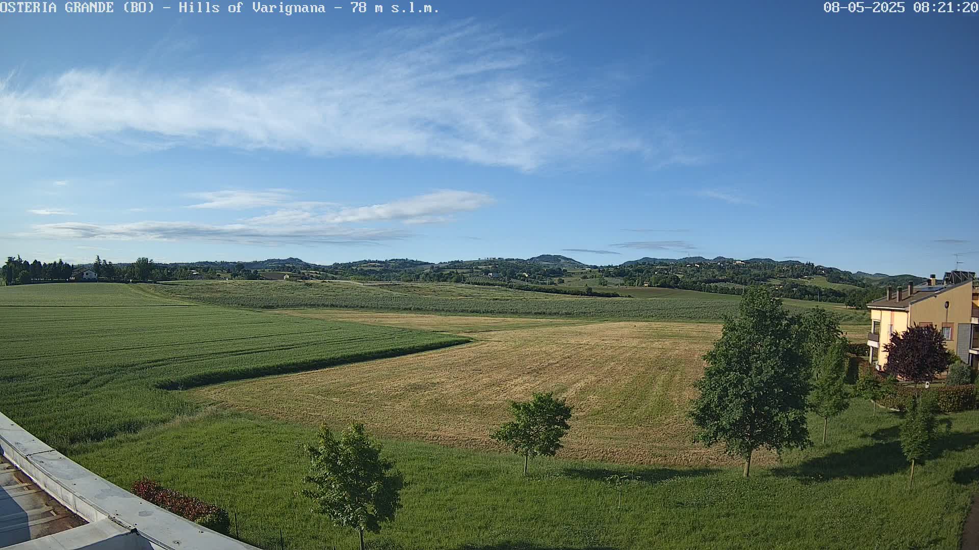 A partly sunny day reveals rolling green and tan fields, a few trees, and a building in the distance, all set against a backdrop of low hills under a mostly clear sky.