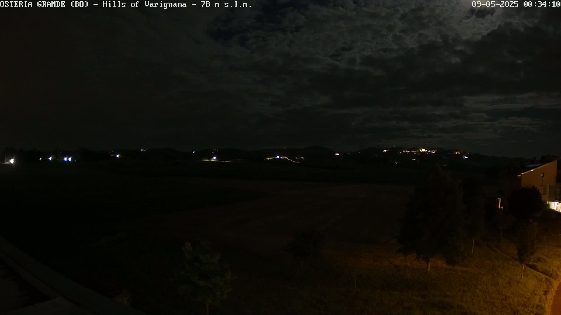A nighttime view of dark hills in the distance, sparsely lit with small lights, under a cloudy sky.