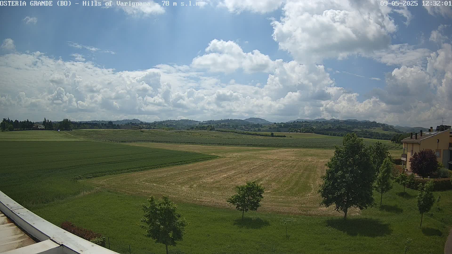 A partly cloudy sky overlooks a wide expanse of farmland, with green fields, some harvested, and distant hills in the background.