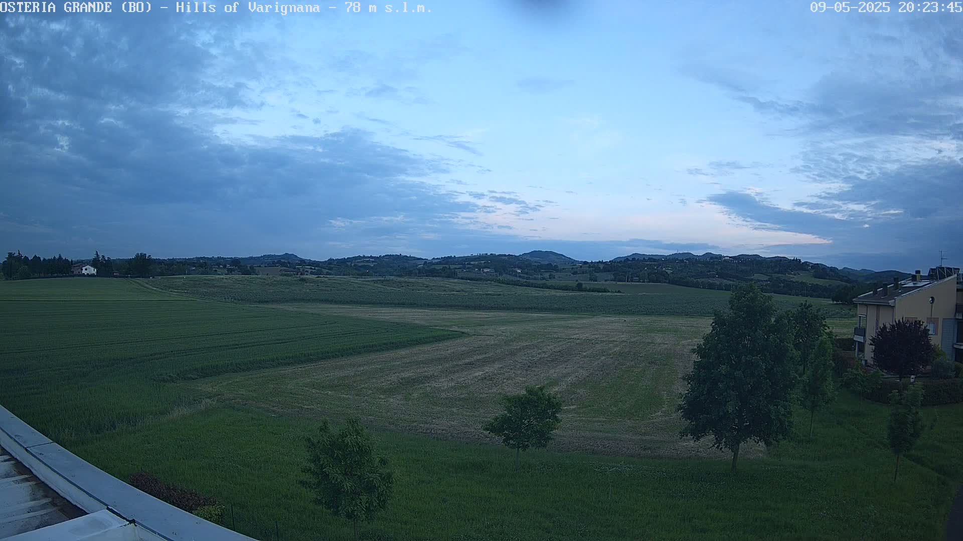 A wide, expansive view of rolling green hills and fields under a mostly cloudy, twilight sky, with a few trees and buildings visible in the foreground and background.