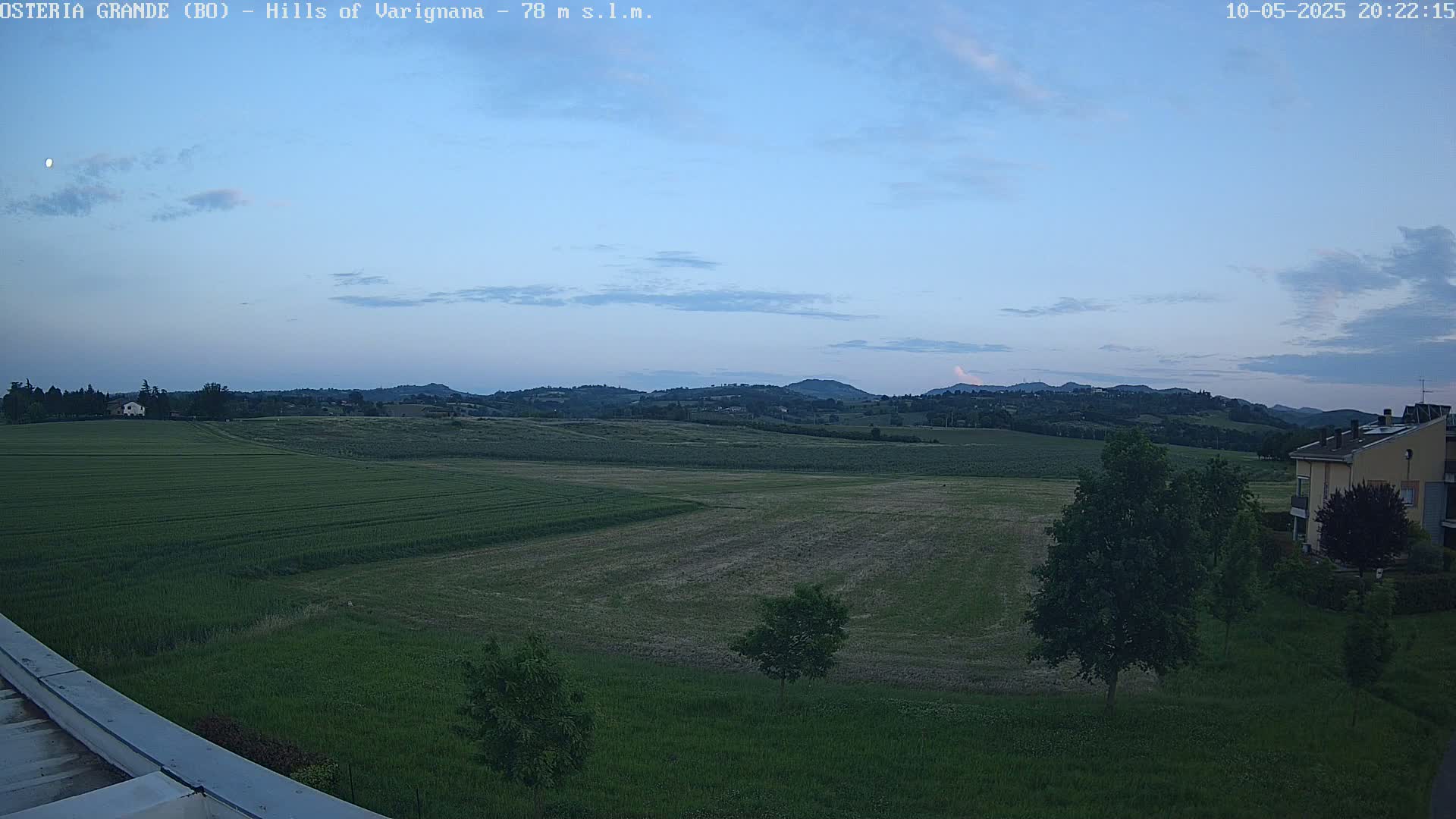 Under a twilight sky with a few clouds, a wide expanse of green fields stretches towards rolling hills in the distance, with a building visible near the lower right.