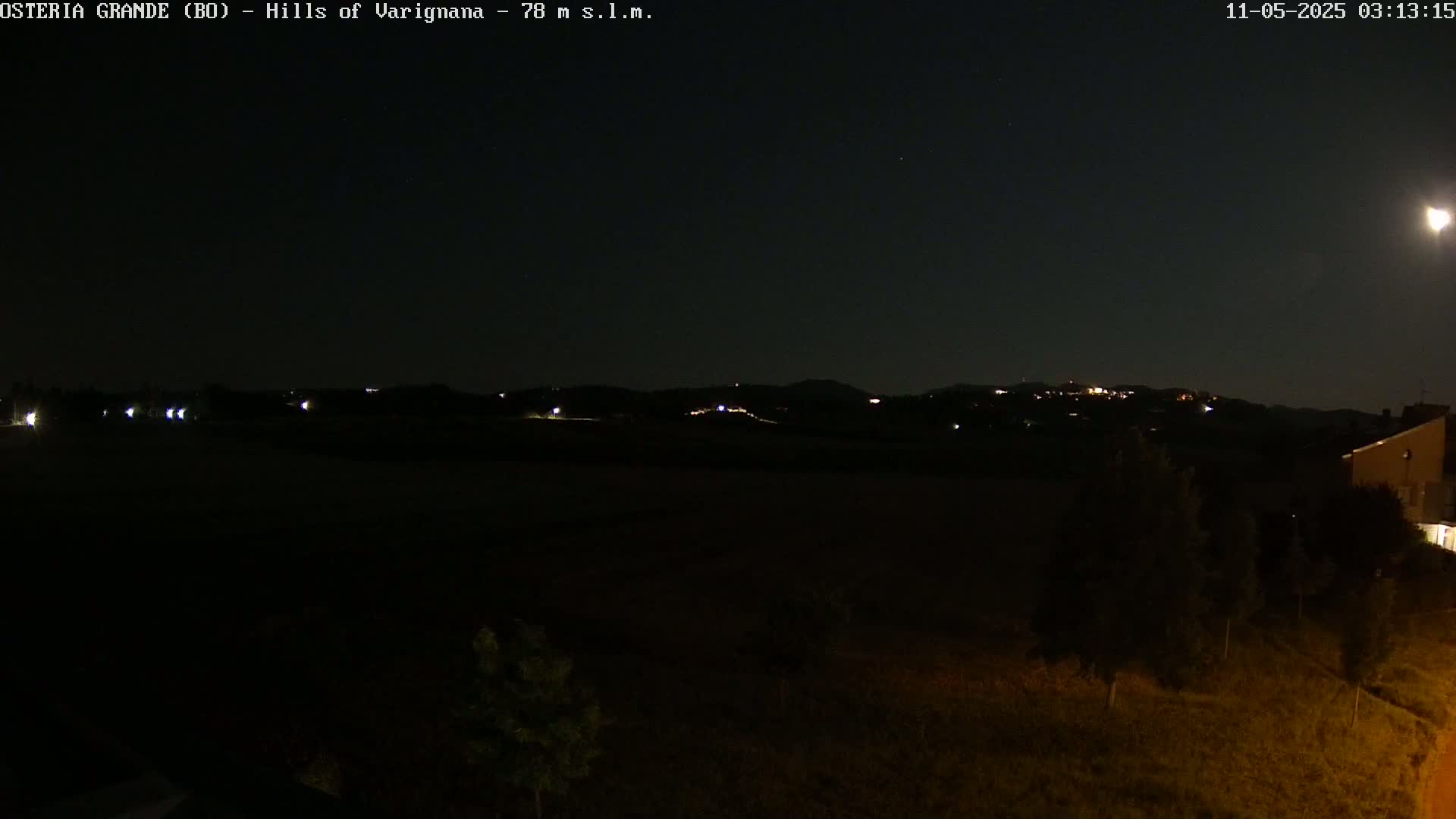 A nighttime view of dimly lit hills in the distance, with a few lights visible in the foreground and a mostly clear sky.