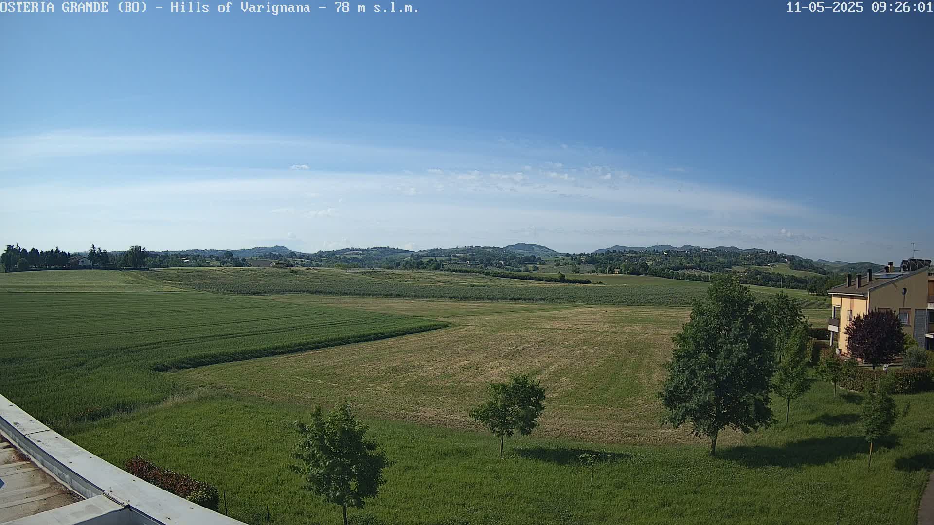 A mostly sunny day reveals a wide expanse of verdant fields, dotted with trees and a building in the distance, all set against a backdrop of rolling hills under a clear blue sky.