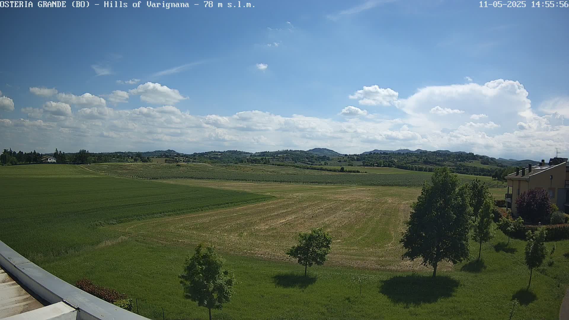 A partly cloudy sky overlooks a flat expanse of green and brown farmland, dotted with trees and a building in the distance.