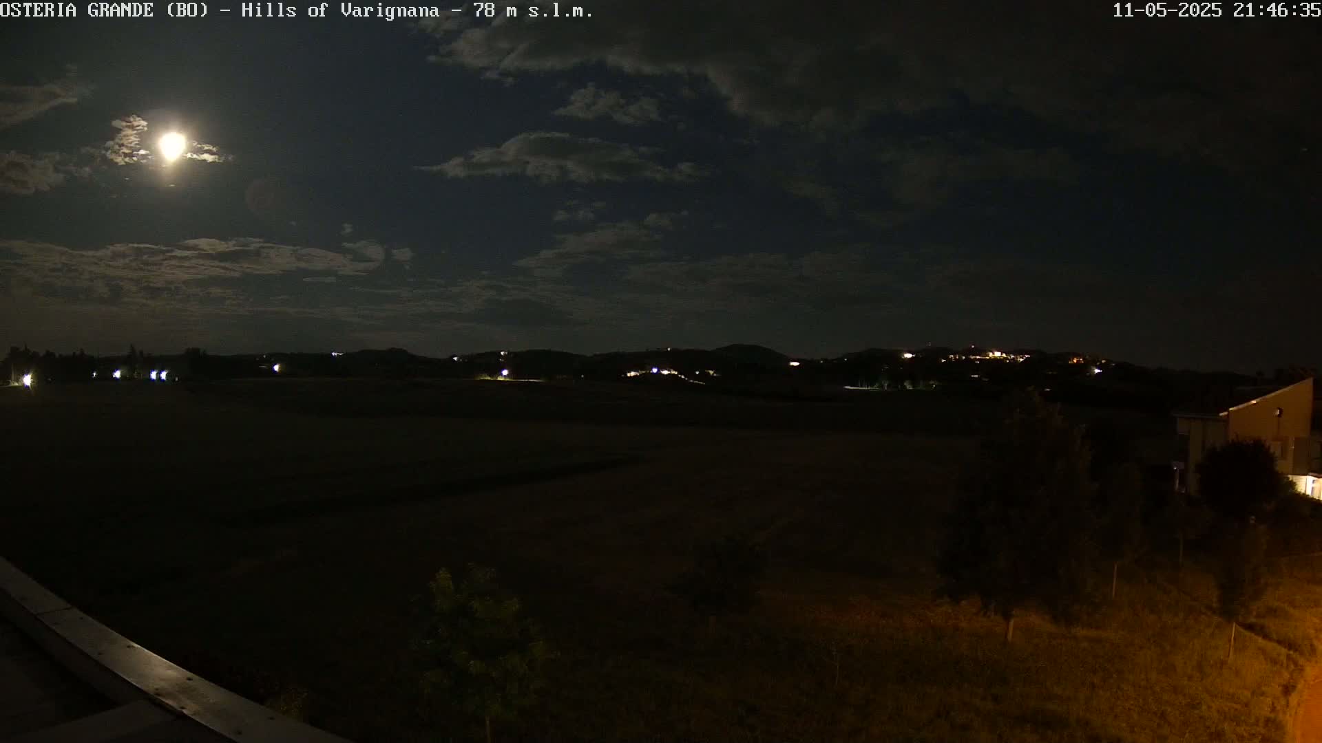 A bright moon illuminates a dark, nighttime landscape of rolling hills with scattered lights, partially obscured by clouds.