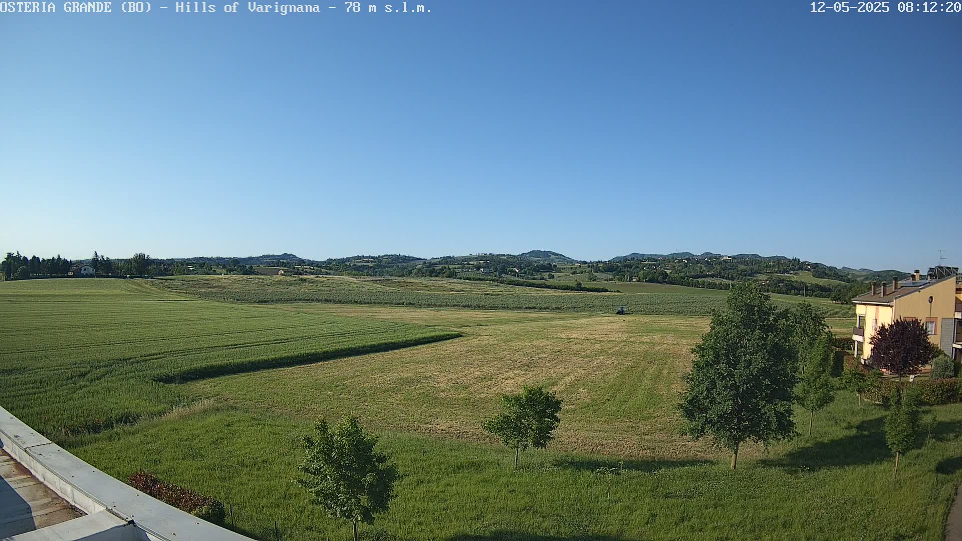 Under a clear blue sky, a vast expanse of farmland stretches out, dotted with a few trees and a building in the distance.