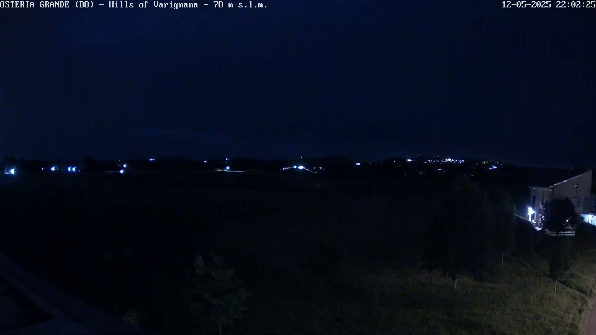 A nighttime, dark view of a hilly landscape with sparsely spaced lights visible in the distance and a building to the lower right.
