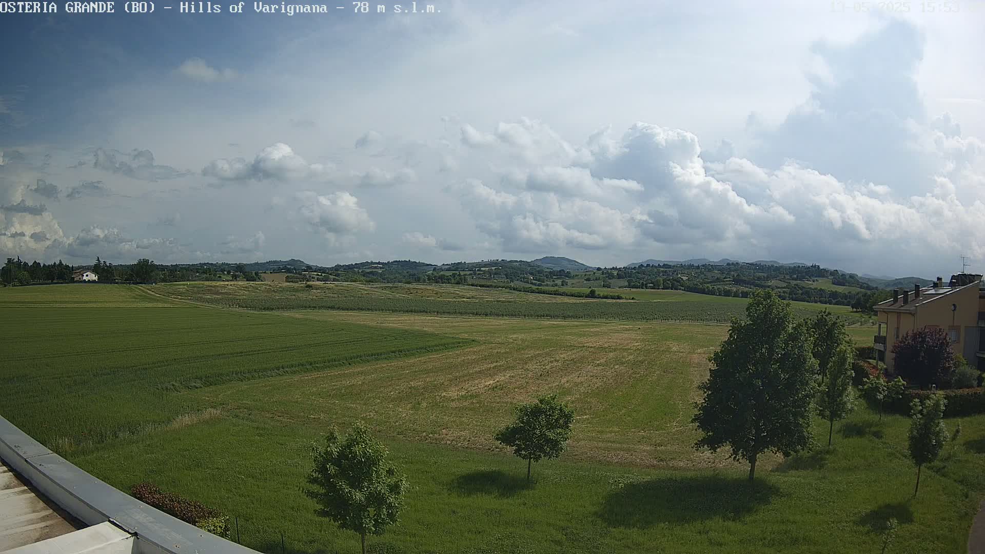 A partly cloudy sky overlooks a vast, green field dotted with trees and a few buildings in the distance.
