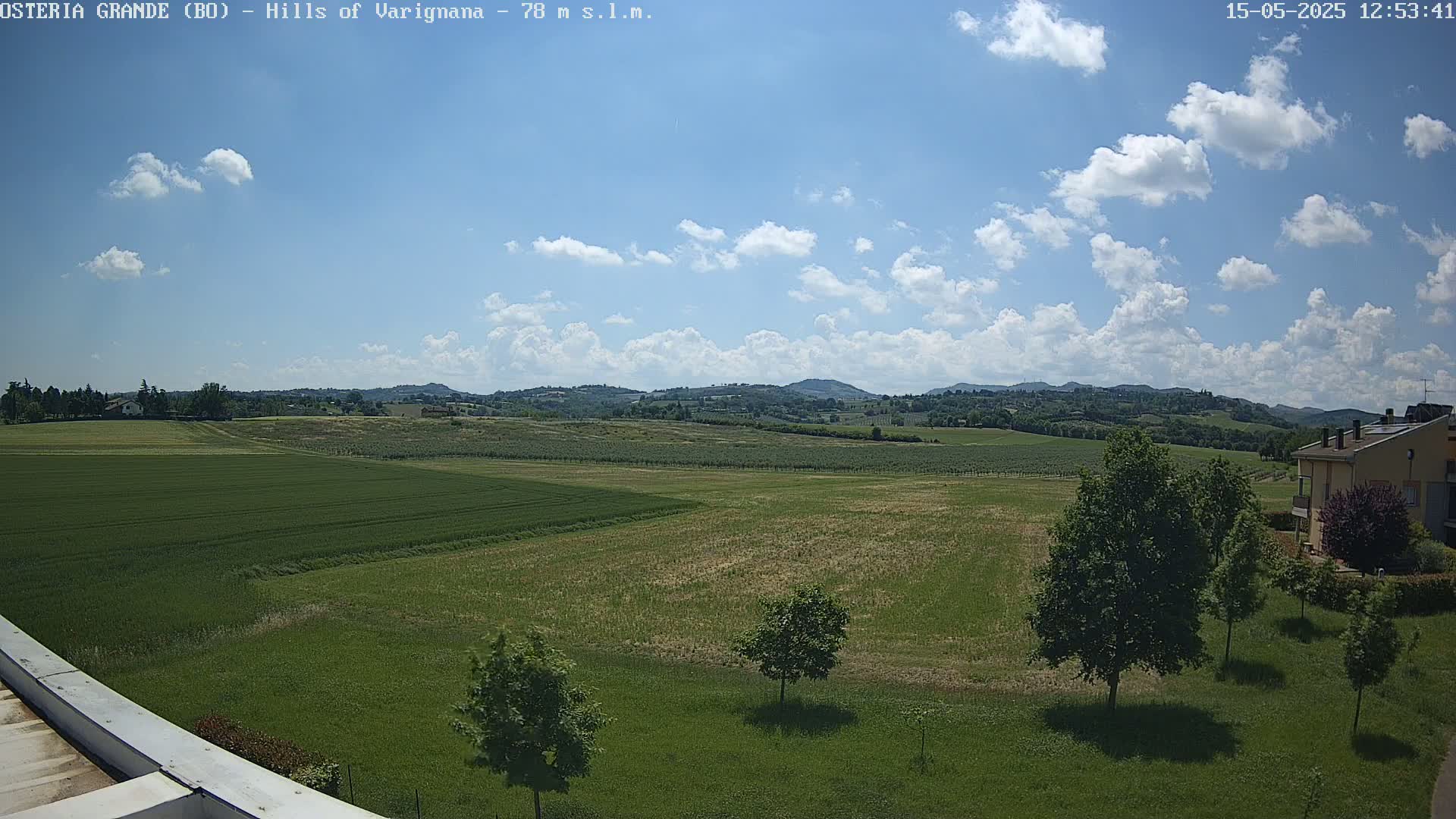 A partly cloudy sky overlooks expansive green fields and distant rolling hills, with a few trees and a building visible in the foreground.
