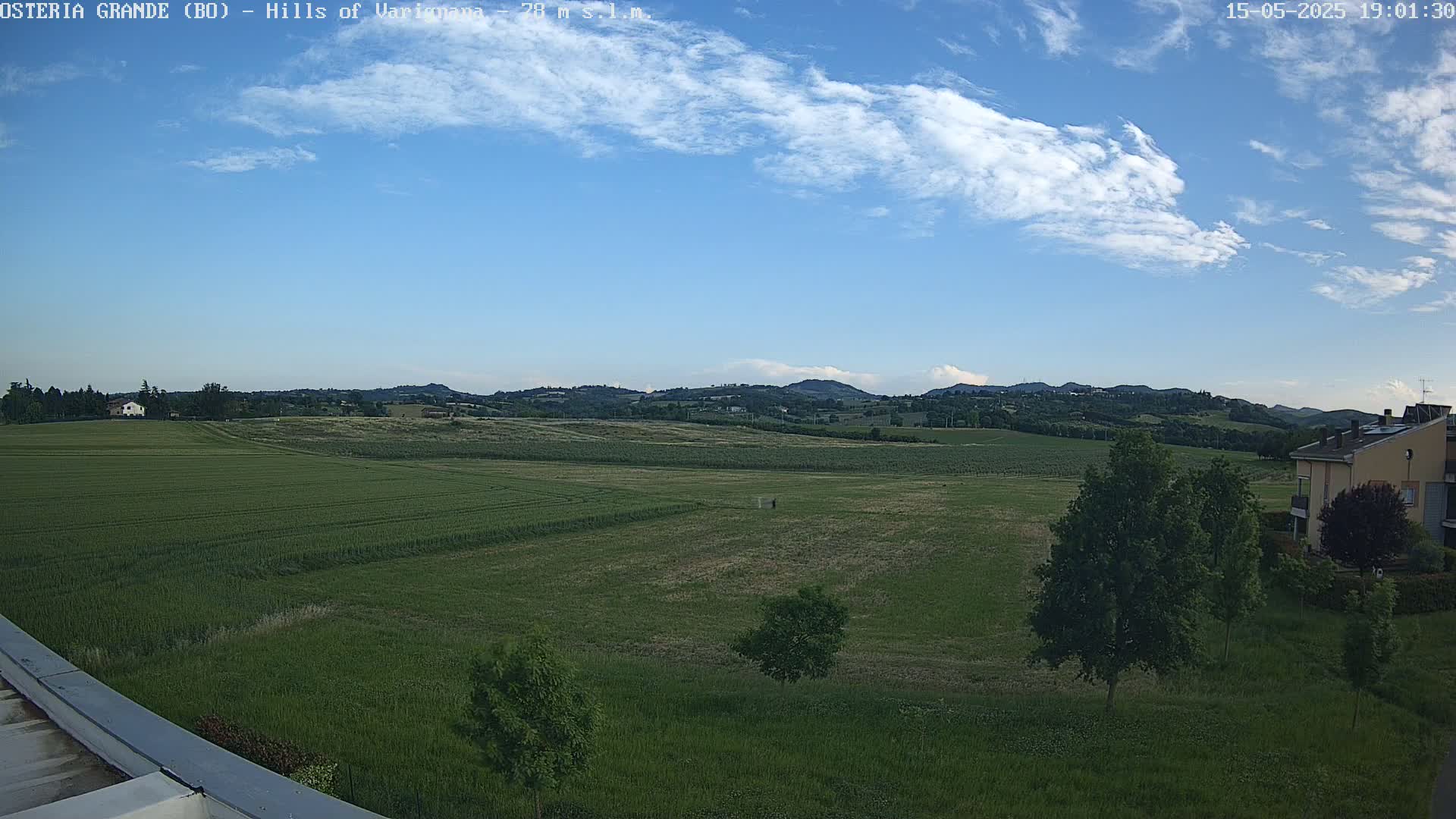 A partly cloudy blue sky overlooks expansive green fields and distant hills, with a few trees and buildings visible in the foreground.