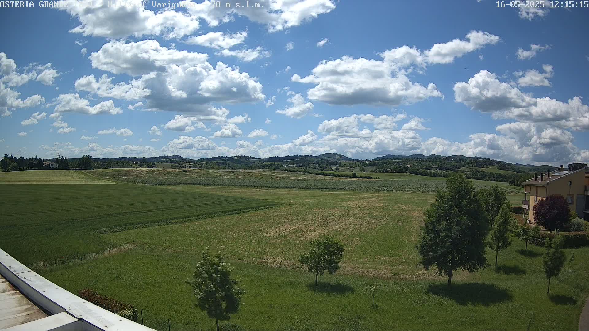 A partly cloudy sky overlooks expansive green fields, a few trees, and a distant residential building under a sunny sky.