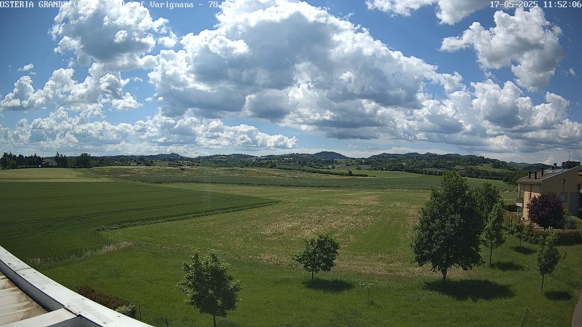 A partly sunny day reveals a sprawling landscape of green fields, dotted with trees and a distant building, under a sky filled with fluffy cumulus clouds.