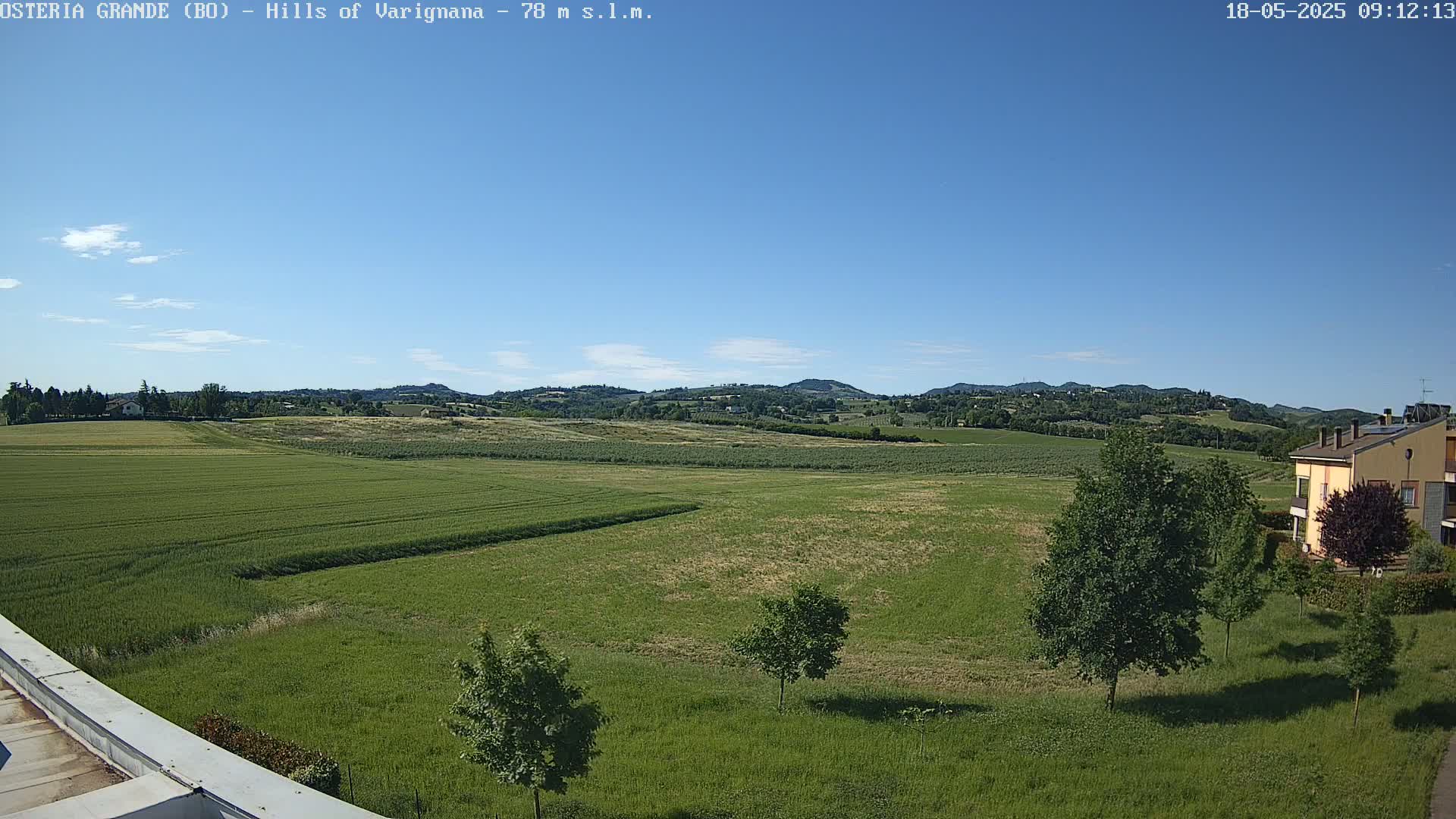 Under a clear blue sky, a vast expanse of green fields stretches toward rolling hills in the distance, with a few trees and a building visible in the foreground.