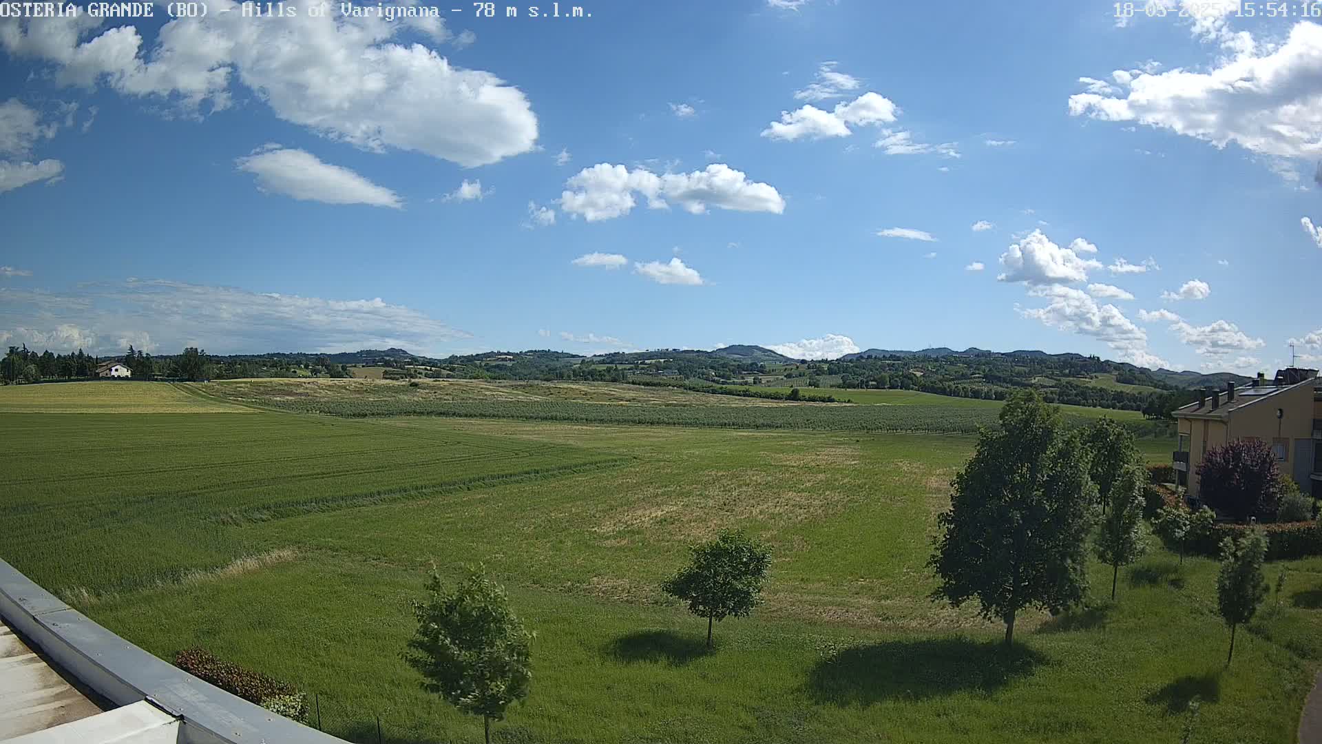 A mostly sunny day with scattered clouds shows a wide expanse of green fields, a few trees, and distant rolling hills.