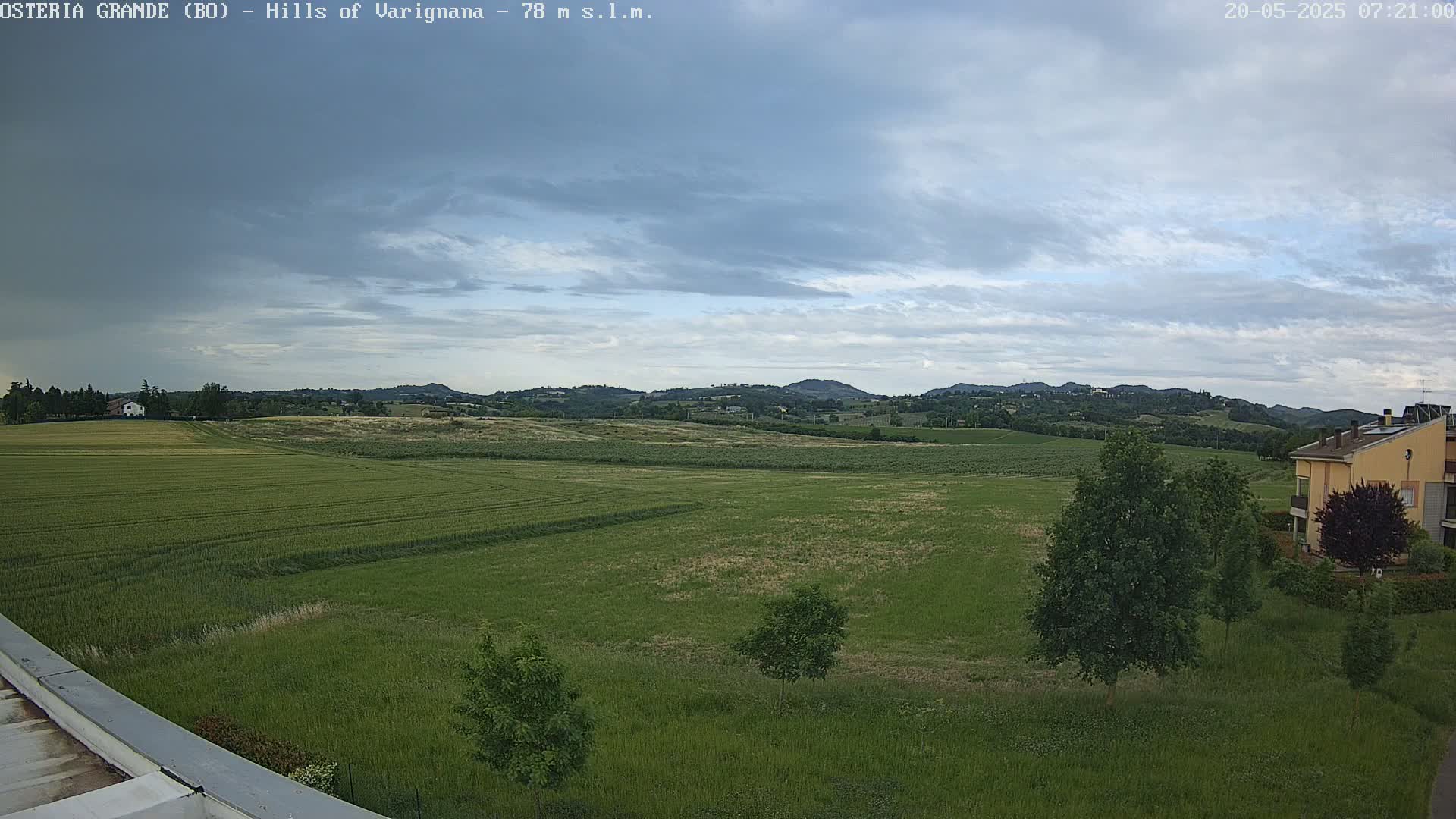 A wide, expansive view of rolling green fields under a mostly cloudy sky shows distant hills in the background and a few buildings near the bottom right.