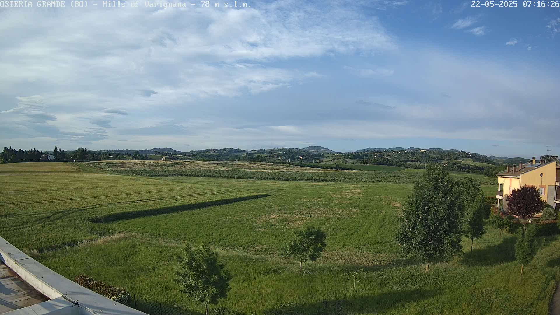 A partly cloudy sky overlooks a vast expanse of green fields and distant rolling hills, with a few trees and a building visible in the foreground.