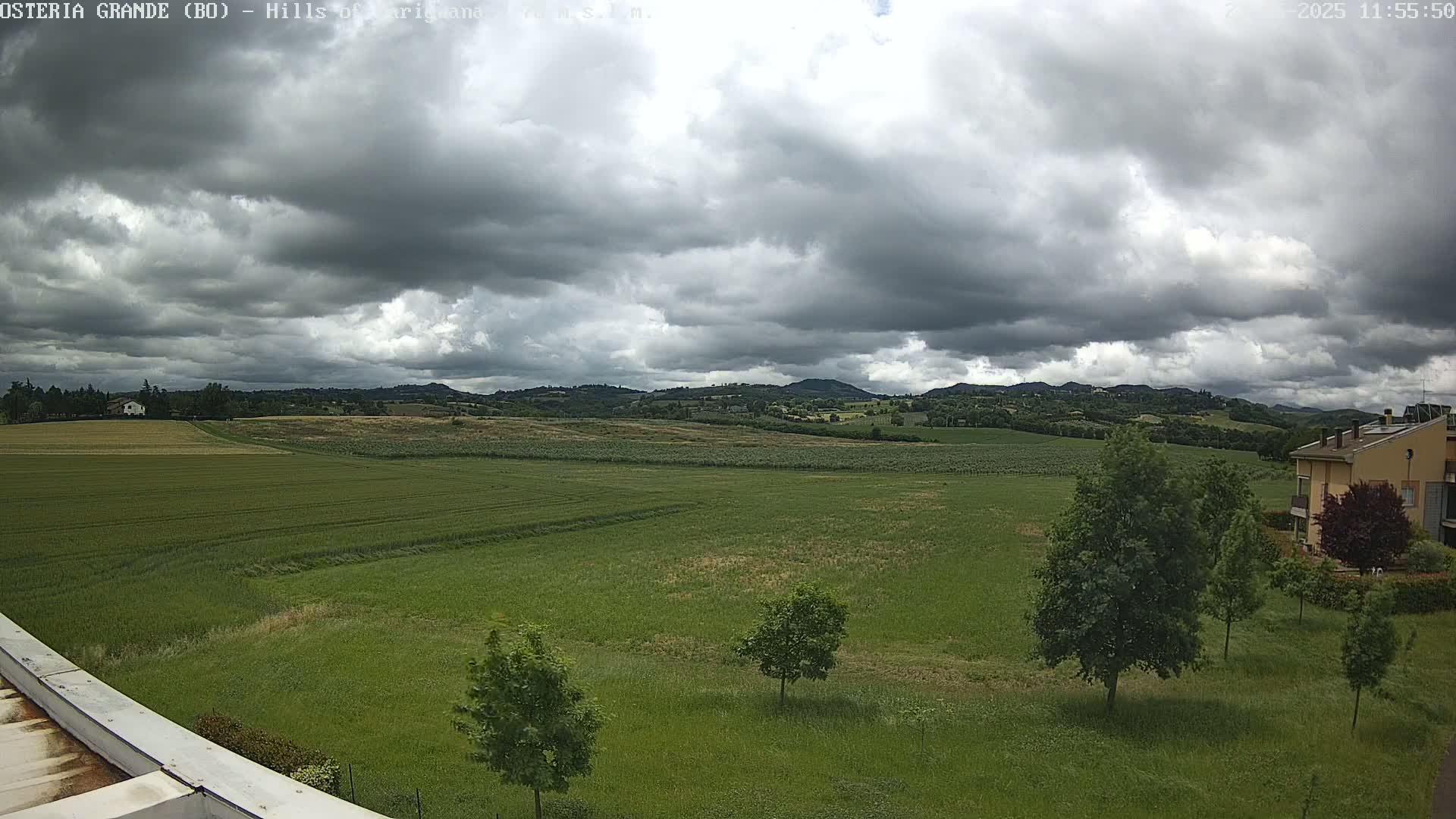 A wide, green field stretches out beneath a cloudy sky, with distant hills visible in the background and a few trees and buildings scattered around.