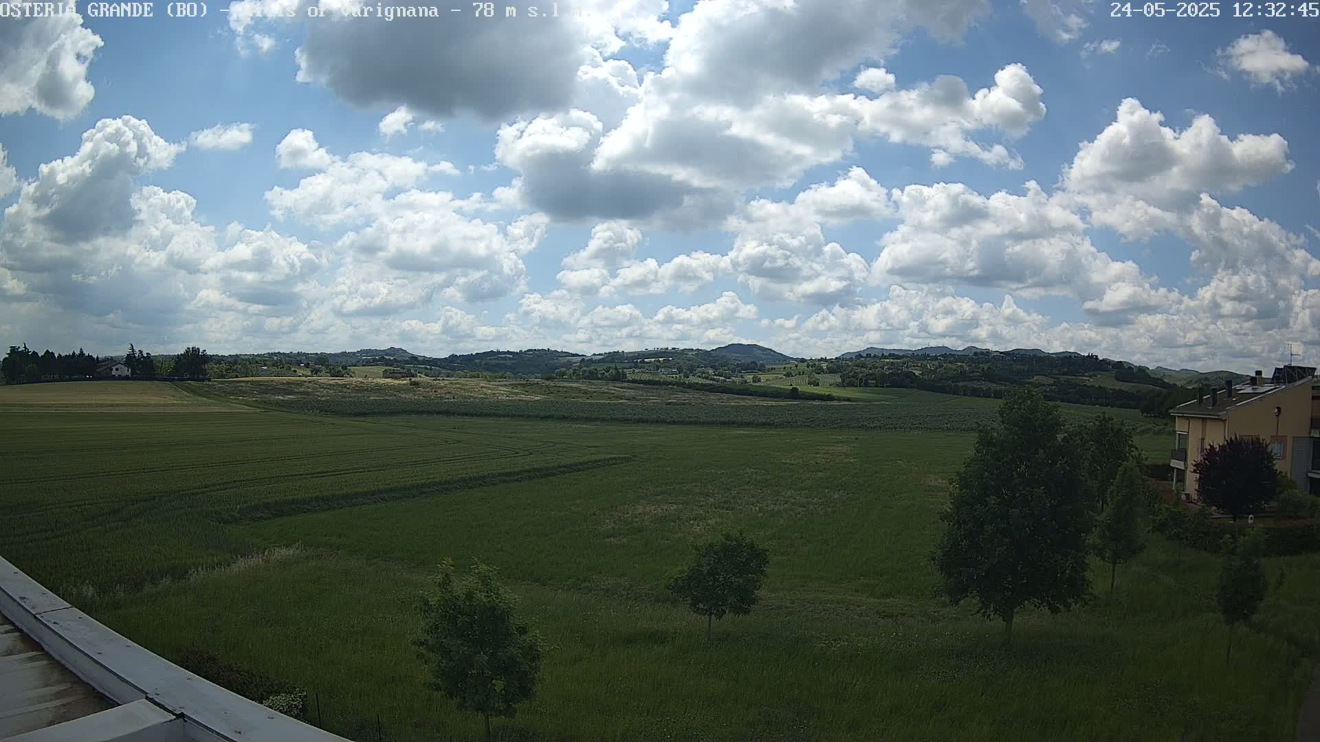 A partly cloudy day reveals a wide expanse of green farmland, dotted with trees, extending to rolling hills in the distance.