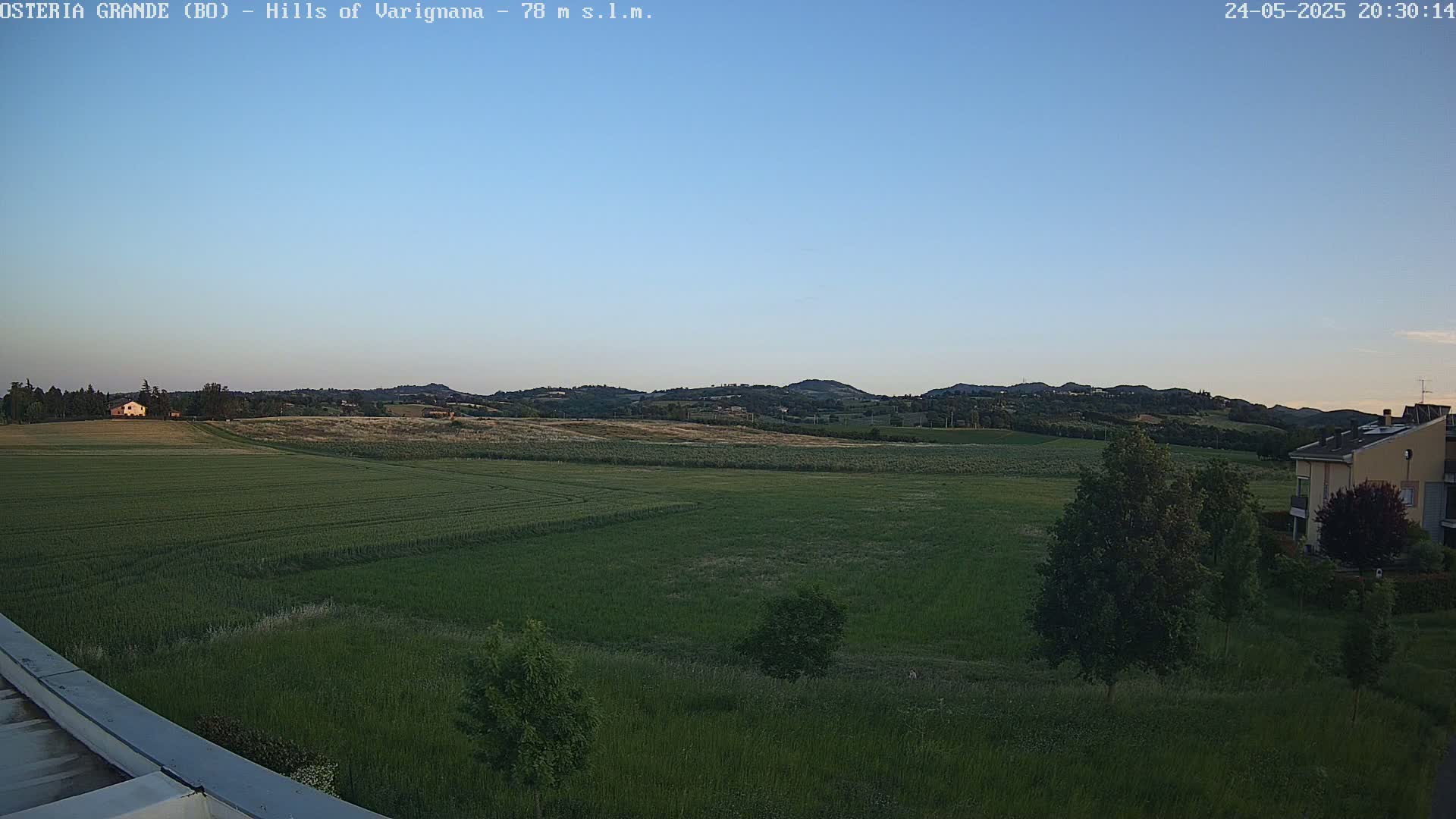 A wide, expansive view of a verdant field under a clear, twilight sky, with distant hills and a few buildings visible in the background.