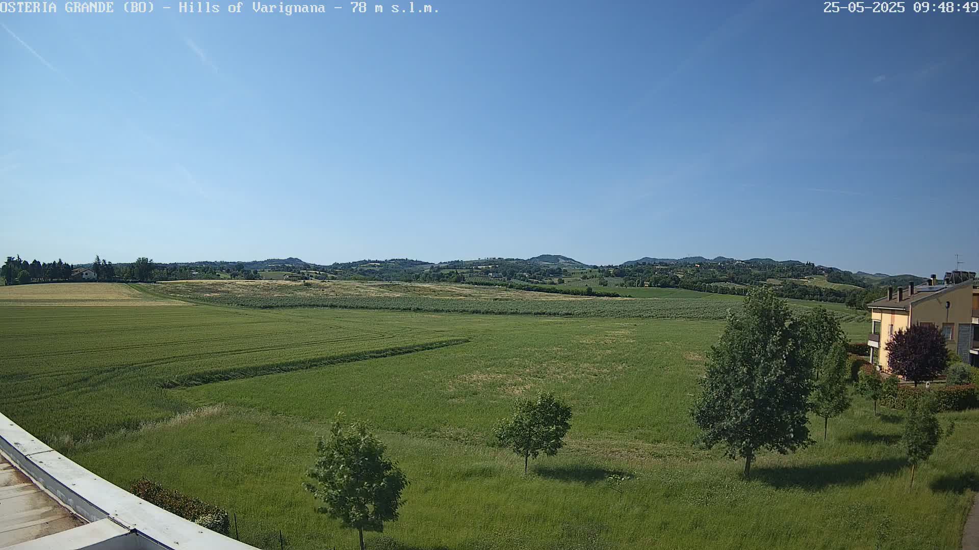 A wide, expansive view of verdant fields under a clear blue sky, with distant rolling hills and a few buildings visible in the lower right corner.